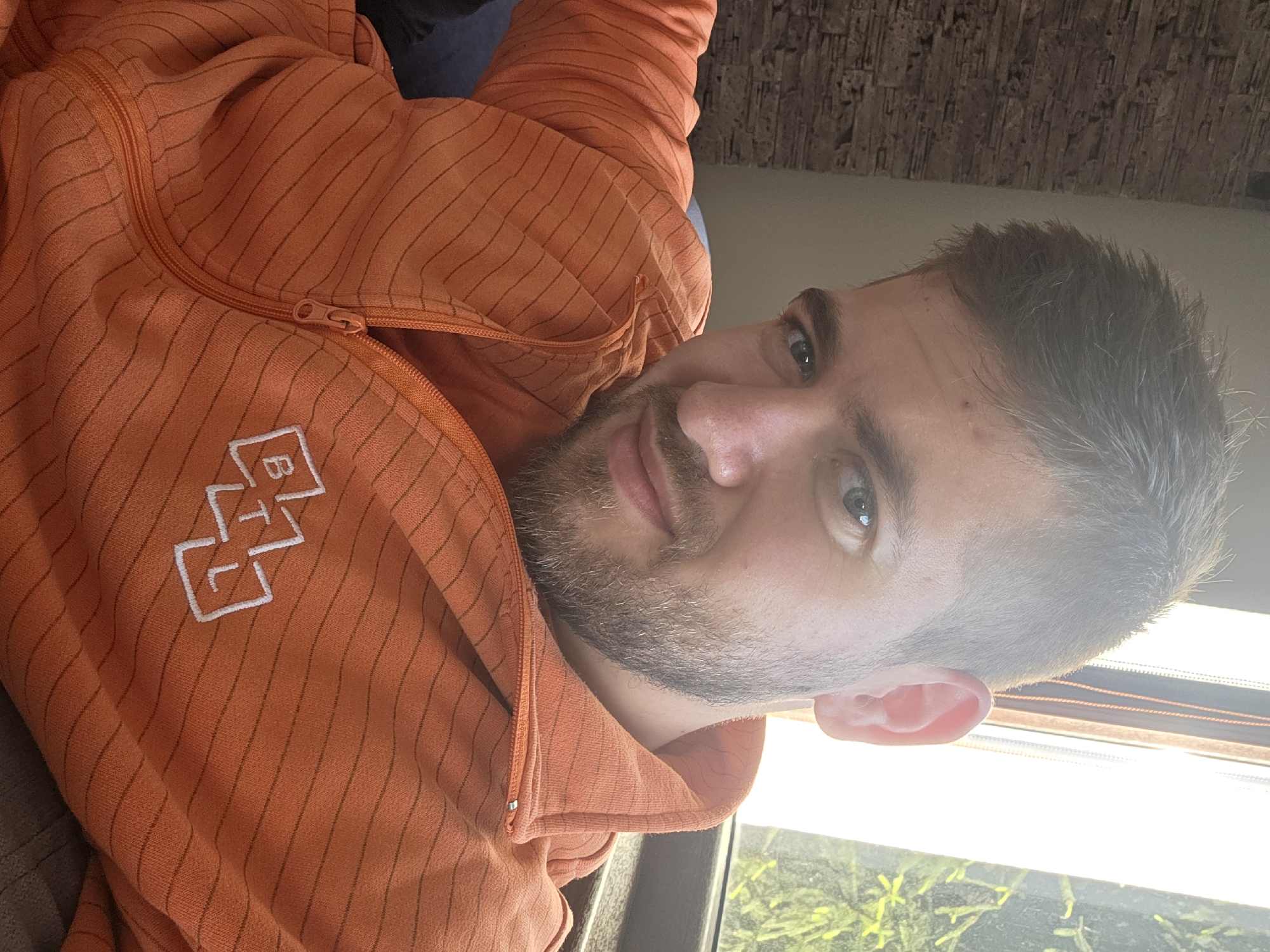 A close-up selfie shows a man wearing an orange zippered hoodie with a white logo on the left breast. The man has short, light brown hair, a beard, and light eyes. The background is partially visible, with a textured wall behind the man and a window showing greenery outside. The setting could be Yagodovo, Bulgaria, but no specific information in the image confirms this location.