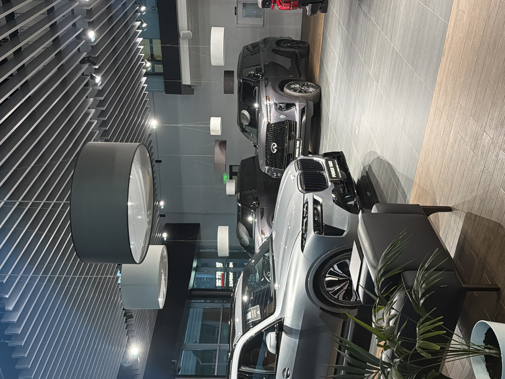 An interior view of a car dealership showcases several luxury vehicles. In the foreground, a silver SUV with the "POWER YARD" badge is prominently displayed. To its right, a dark gray Infiniti SUV also features the "POWER YARD" badging on its front. In the background, another SUV appears to be partially visible. The showroom features a modern design with a slatted ceiling, recessed lighting, and large pendant lamps. The flooring is a combination of large gray tiles and wood-look planks. A dark gray leather armchair and a potted plant are positioned in the left foreground, adding to the sophisticated ambiance of the car dealership.