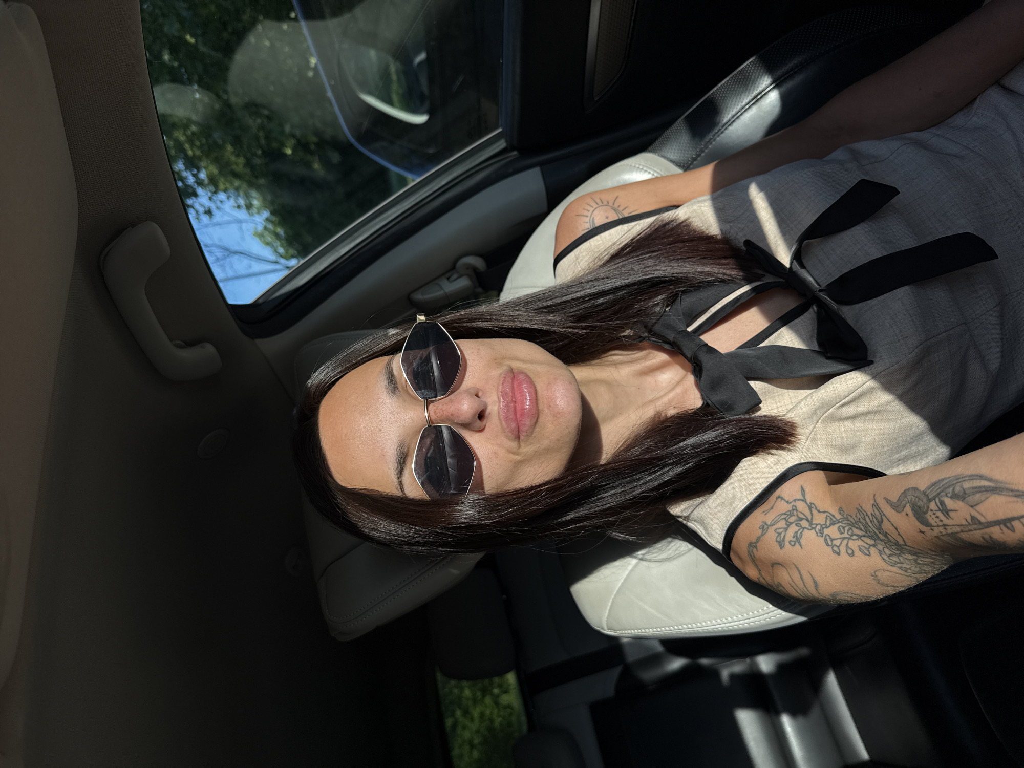 The image captures a vertically oriented selfie of a woman inside a car, taken during bright daylight, likely in Pleven, Bulgaria. The woman, positioned in the upper portion of the frame, has long dark hair and wears sunglasses that reflect the surrounding light. Her lips are slightly parted, and she holds a neutral expression. She is dressed in a light-colored top, possibly beige, featuring a prominent black bow or tie detail at the collar. A small sun tattoo is visible on her right arm, while her left arm displays a larger, more detailed tattoo with floral and abstract designs.
She is seated in a light-colored leather car seat, with parts of the dark car interior visible around her. Strong, diagonal shafts of sunlight cut across the scene, creating distinct patterns of light and shadow on her clothing and the car's interior. Through the car's sunroof and side windows, a clear blue sky and vibrant green tree foliage are visible, indicating an outdoor setting on a sunny day. A light-colored grab handle is also noticeable on the car's ceiling near the sunroof. The image conveys a casual moment in a well-lit car interior. No visible text is present.