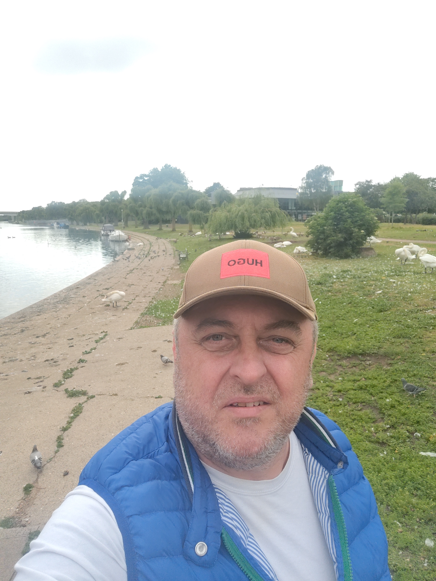 A selfie-style photograph captures a middle-aged male with light facial hair, wearing a brown baseball cap with an upside-down red "HUGO" logo, a white long-sleeved shirt, and a blue quilted vest. The scene is set outdoors along the River Nene in Peterborough, United Kingdom, specifically in the vicinity of the Embankment Park and the Key Theatre, the latter being discernible in the background behind a line of trees.
To the subject's left, a concrete pathway borders the river, where several small boats are moored. Numerous pigeons are scattered across the pathway, and a single white swan stands on the concrete. To the subject's right, a grassy area, dotted with small white flowers, contains a flock of white birds, primarily swans, resting or foraging. Additional pigeons are visible on the grass. The background features a dense line of various deciduous trees, including willows, obscuring parts of the distant buildings. The sky is overcast and appears bright.