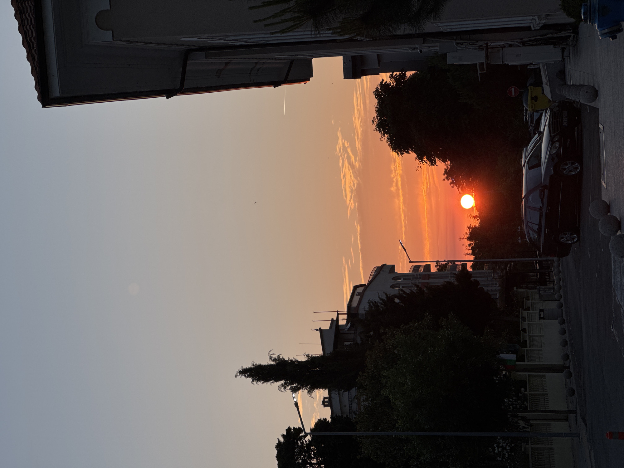 The image depicts a sunset scene in Tsarevo, Bulgaria, captured at street level. The sky transitions from a light blue at the zenith to vibrant orange and red hues near the horizon, where the sun appears as a bright, circular, orange-red orb, partially obscured by silhouetted trees. Scattered, elongated clouds are present in the middle sky, illuminated by the setting sun. A faint airplane contrail is visible in the upper right quadrant, and a subtle circular light source is present in the upper left.
On the left side of the frame, a multi-story building with a light-colored facade and arched windows is partially visible, adorned with balconies. Behind it, another building features a flat roof with antenna structures and a distinct white object, possibly a bird or a statue. In the foreground on the right, a building with a red-tiled roof, light facade, and a visible downspout occupies the edge of the frame.
The street level features a paved road and a sidewalk/parking area demarcated by concrete spherical bollards. A black Mercedes-Benz E-Class wagon, bearing the Bulgarian license plate "CT 7328 DX", is parked on the right side, facing left. A yellow public waste bin is located behind the parked vehicle. On the left, a decorative fence or wall separates the street from properties, displaying a small flag with vertical green, white, and red stripes. A red and white striped bollard or cone is situated in the lower-left corner.
Numerous trees, primarily dark silhouettes against the bright sky, are dispersed throughout the scene, with a particularly tall, slender conifer-like tree prominent on the left. Two streetlights are visible and illuminated: one on the left foreground and another further down the street on the right. No human figures are discernible in the image.