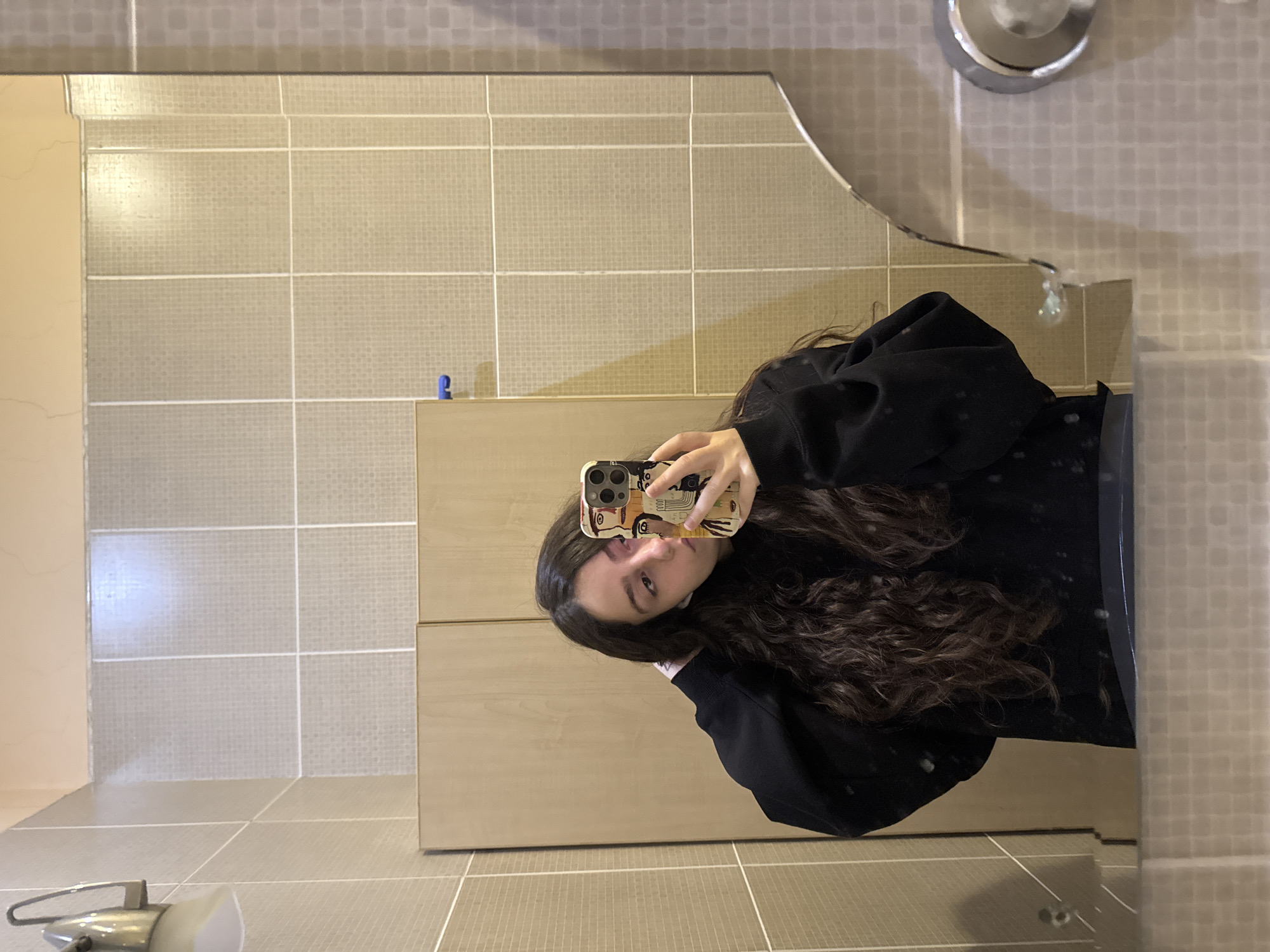 The image is a selfie taken in a bathroom, likely in Yerevan, Armenia, based on the bathroom's style. The person taking the selfie is a young woman with long, wavy brown hair, wearing a black hoodie. She is holding a smartphone with a decorative case. The bathroom has a tile wall with horizontal lines, a light-colored cabinet, a sink, and a chrome faucet. There is a blue object on the cabinet. The reflection shows the woman holding up the phone to take the picture in the mirror.