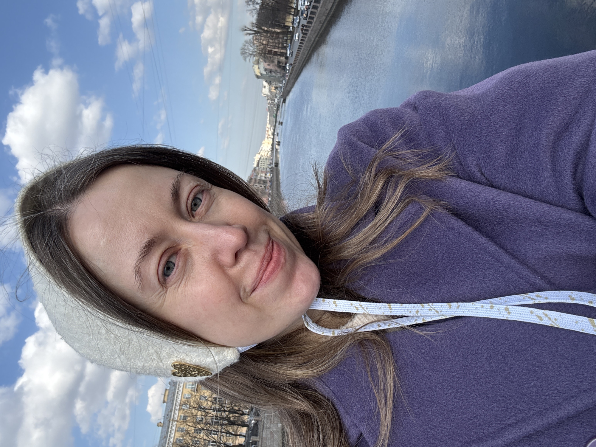 An outdoor selfie captured from a bridge overlooks the Moika River (Moyka River) in Saint Petersburg, Russia. The central subject is a woman, positioned slightly to the right of the frame, looking directly at the camera with a faint smile. She has light brown hair, fair skin, and blue-grey eyes. She wears a white textured bonnet adorned with a gold heart-shaped emblem on her left side, and a purple, possibly wool, coat. A light blue patterned ribbon hangs from the bonnet.
In the background, the Moika River flows, reflecting a bright blue sky with scattered white cumulus clouds. Multiple small boats are visible in the distance on the river's surface. The left bank of the river features a continuous line of multi-story historical buildings, including a prominent light yellow structure with classical architectural details. Bare deciduous trees are visible in front of these buildings, lining a street with parked and moving vehicles. The right bank displays a stone embankment, a road with vehicular traffic, and additional buildings partially obscured by bare trees. The scene is illuminated by natural light, indicative of daytime.