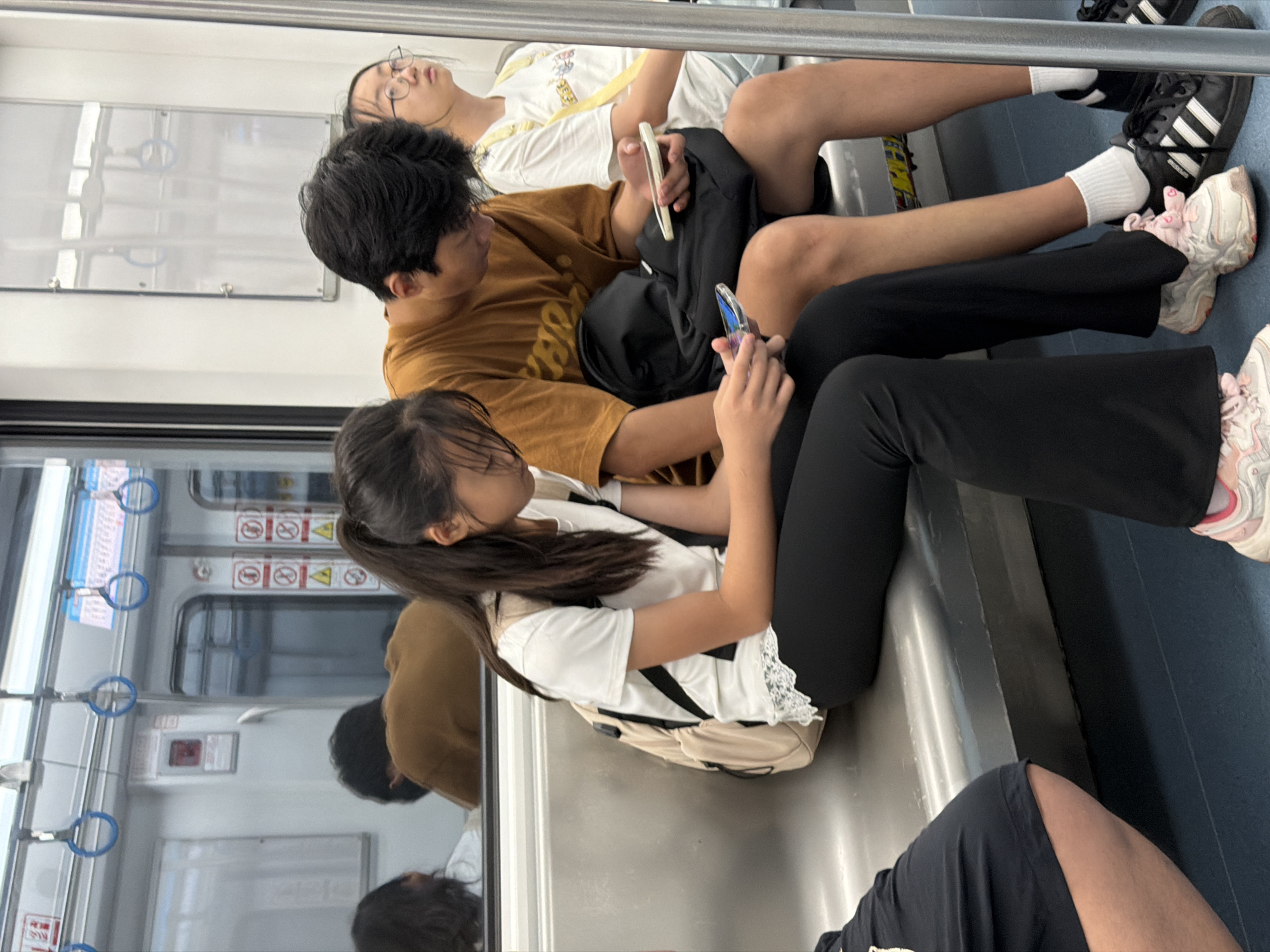 In the unknown, unknown city, on a subway train, three young people are seated. The person in the foreground on the left, a female, is wearing black pants and a white t-shirt with lace trim, and a beige backpack. She is looking down at her phone. To her right, a male wearing a tan t-shirt and black shorts is looking down at his phone, with a black backpack on his lap. To his right, a female wearing a white t-shirt and a yellow strap is looking upwards. The train car is clean with metallic seats and poles for passengers to hold onto. The lighting appears to be artificial.