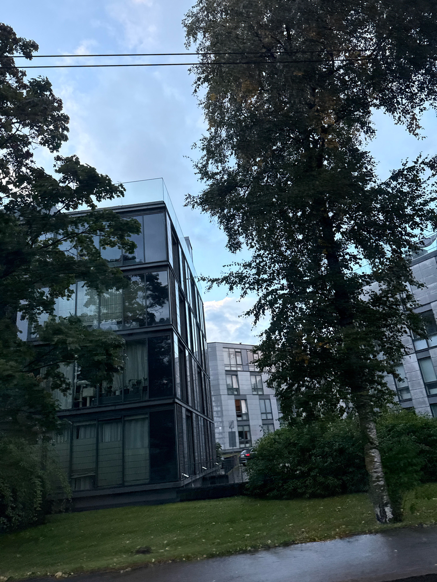The image depicts a modern, multi-story building with a glass facade in Tallinn, Estonia. Trees with dense foliage partially obscure the building, and some leaves appear to be changing color, suggesting it might be autumn. The sky is overcast with some visible clouds. In the foreground, a grassy area and a wet paved surface, possibly a sidewalk or road, are visible, indicating recent rainfall. Power lines run horizontally across the upper portion of the frame. Another residential building with a more traditional facade is partially visible in the background. The scene appears to be residential or mixed-use.