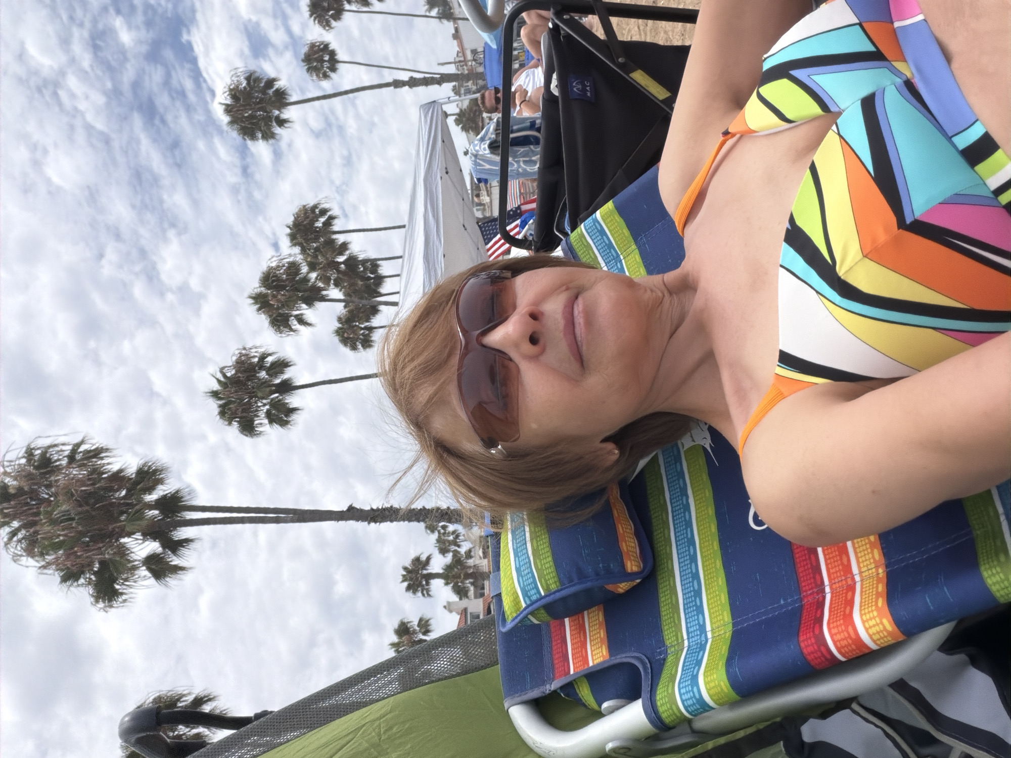 A medium shot captures a woman in a colorful bikini resting in a beach chair. She wears sunglasses and her short, brown hair is windswept. The beach chair, adorned with bright stripes of blue, green, orange, and yellow, is positioned on the sand. In the background, palm trees sway under a cloudy sky. A white canopy tent and American flags are visible further down the beach, where other beachgoers are present. The scene is set at the beach in San Clemente, California.