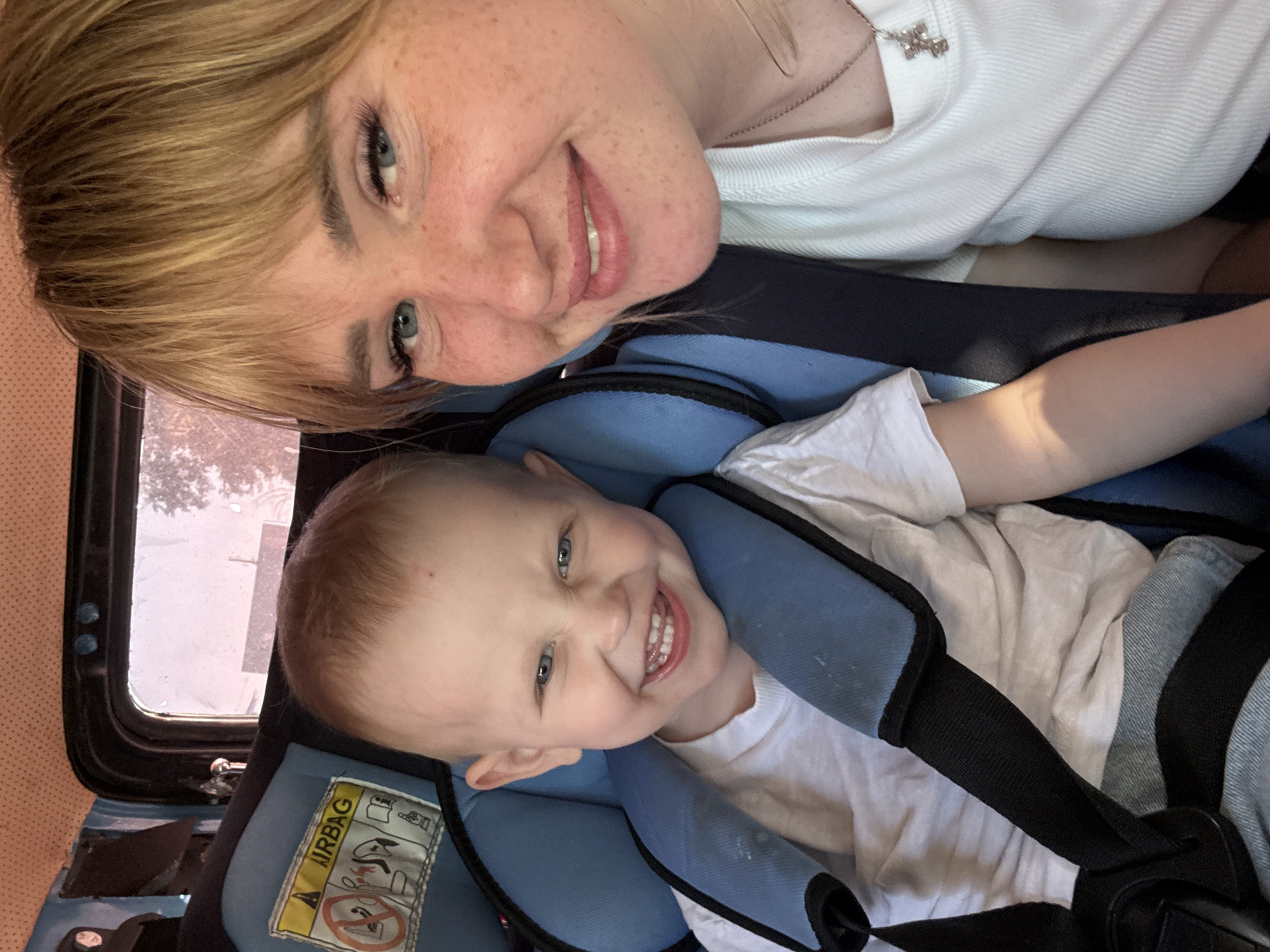 A photograph shows a woman and a young child in a car. The woman, with blonde hair and freckles, is smiling at the camera. She is wearing a white shirt and a necklace with a cross. The child, with blonde hair and blue eyes, is smiling and strapped into a blue car seat. He is also wearing a white shirt. The car seat is positioned near a car window. A sticker with the word "Airbag" and pictograms is visible on the side of the car seat. The location of the image is in the city of Chaikovsky, Russia.
