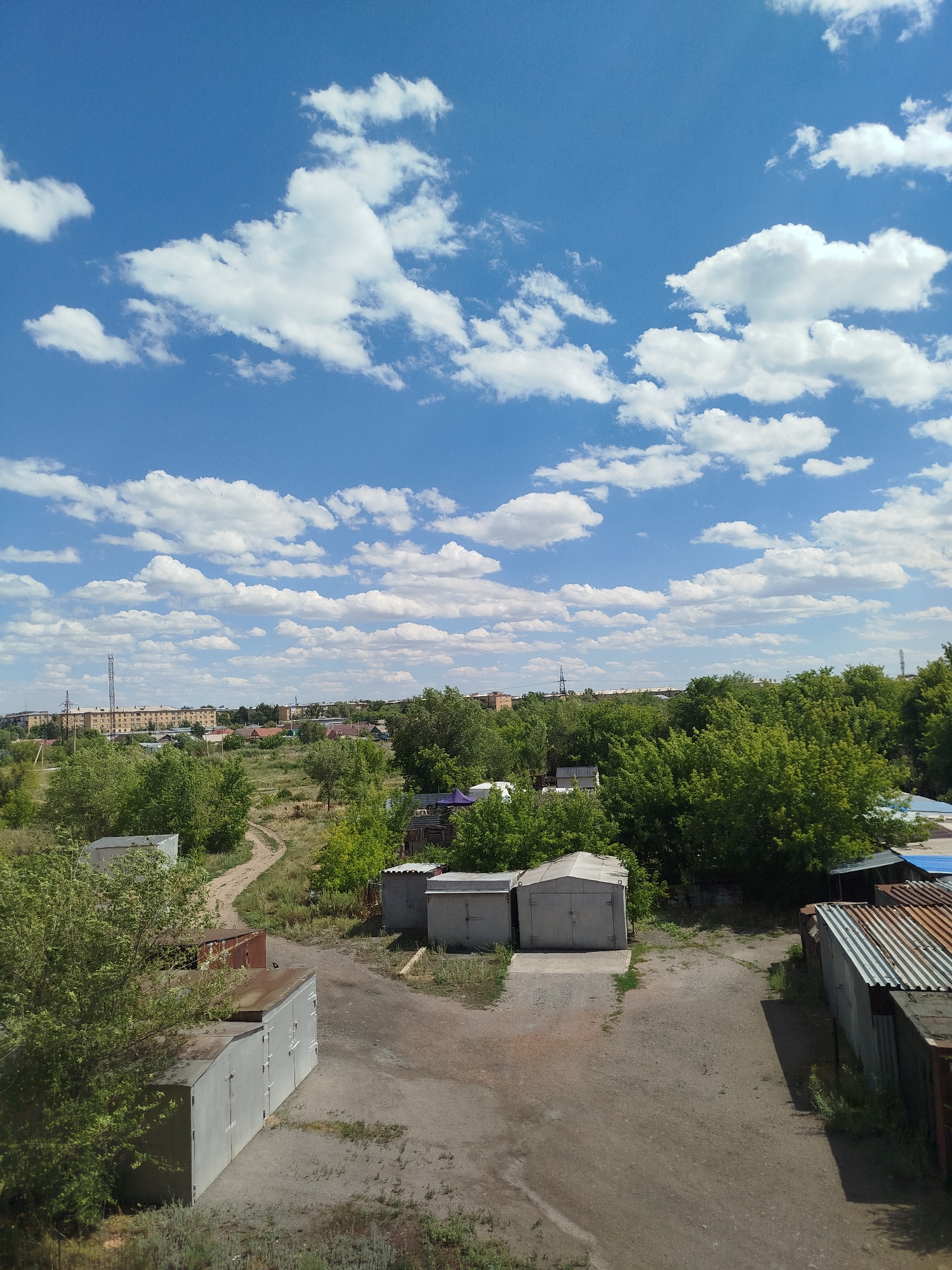 A medium-angle outdoor daylight shot captures a scene of garages and vegetation under a bright blue sky dotted with white cumulus clouds. In the foreground, several gray, rectangular garages with large doors are visible, some appearing weathered and rusted. A dirt path winds through the scene, leading past the garages and into an area with more trees and shrubs. In the background, a small cluster of buildings, including what appear to be apartment blocks and houses, are nestled amongst a dense expanse of green trees. A tall telecommunication tower is also visible in the distance. The overall impression is one of a semi-urban or suburban landscape on a clear day.