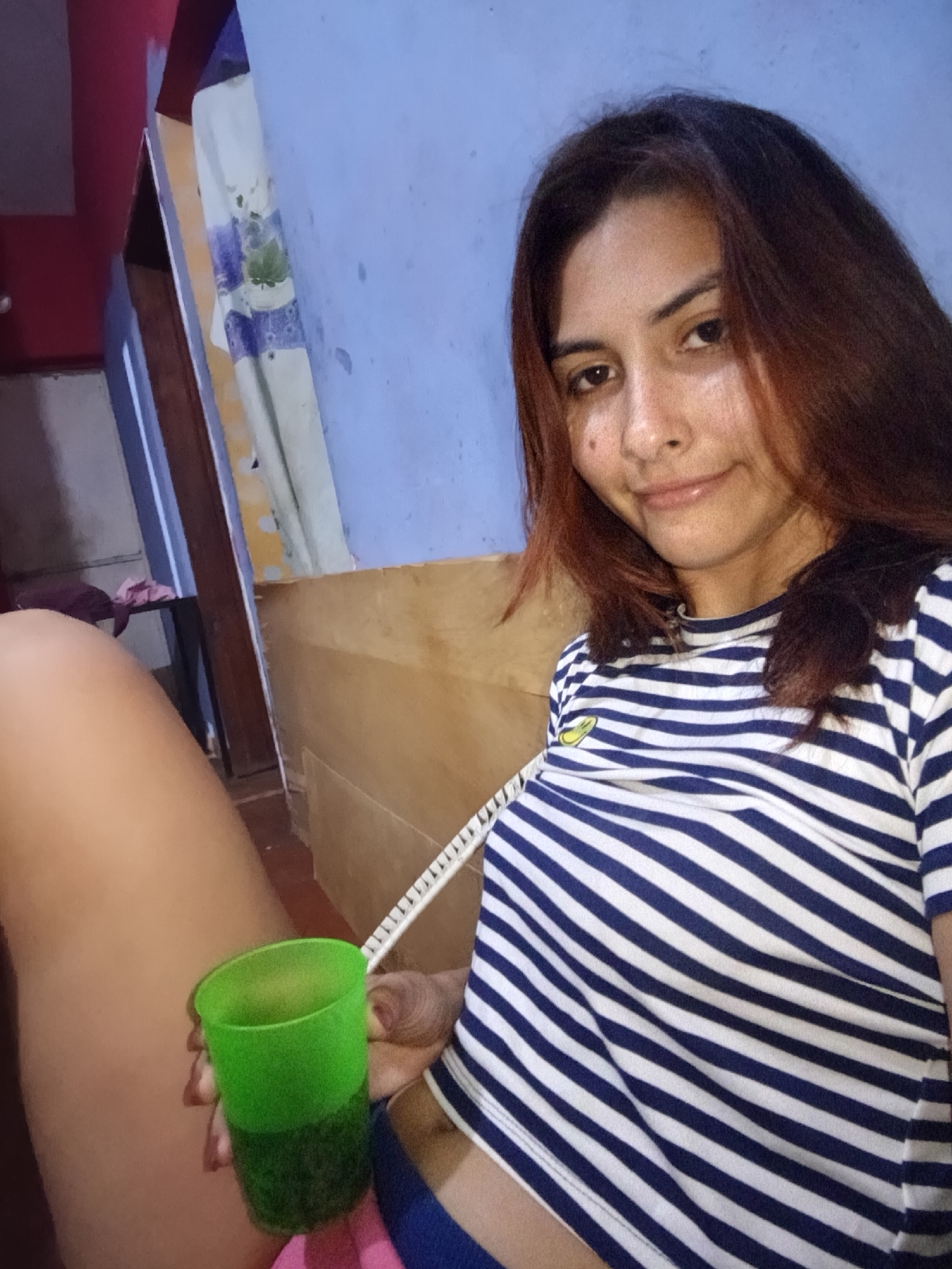 A fair-skinned person with red-tinged brown hair looks at the camera with a slight smile. They are wearing a blue and white striped shirt. In their left hand, they hold a green cup filled with a dark liquid. The person is seated, with a portion of their leg visible. Behind them is a light blue wall, a wooden headboard, and a doorframe with a curtain. No landmarks indicate the city of Juan Griego, Venezuela.
