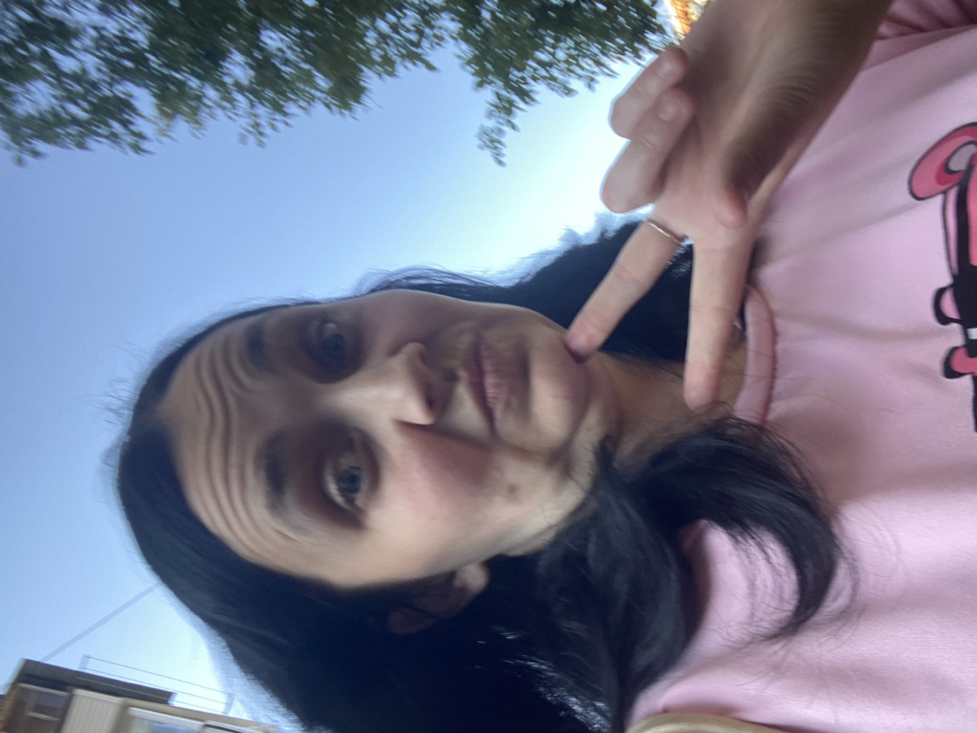 A slightly low-angle, waist-up selfie shows a woman with dark hair and fair skin looking directly at the camera. She is wearing a pink sweatshirt with a black and pink graphic design. Her right hand is raised, forming a peace sign with her index and middle fingers, while her left hand rests on her chin. Her hair is long and dark, framing her face. The background is a clear blue sky with a blurry green tree on the right and the side of a building on the left. The lighting suggests it is daytime.
