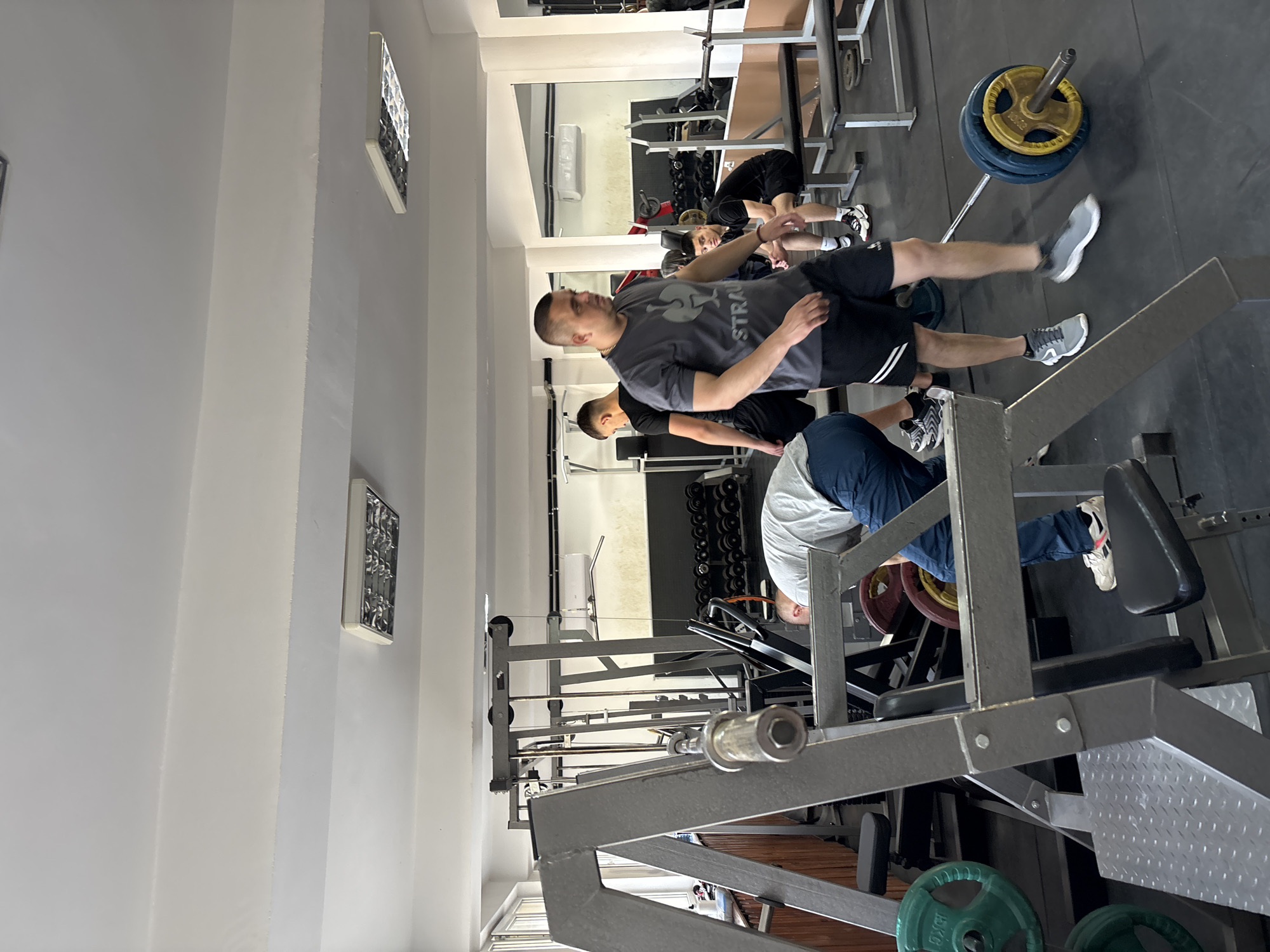 The image shows a brightly lit gym in Zlatograd, Bulgaria. Three individuals are present. One man wearing a gray t-shirt and black shorts is standing. Another man is seated on a leg press machine, wearing a light-colored shirt and blue pants. A third person is lifting weights in the background. Weightlifting equipment, including a barbell with plates, is scattered throughout the gym. The walls are white, with framed decorations.