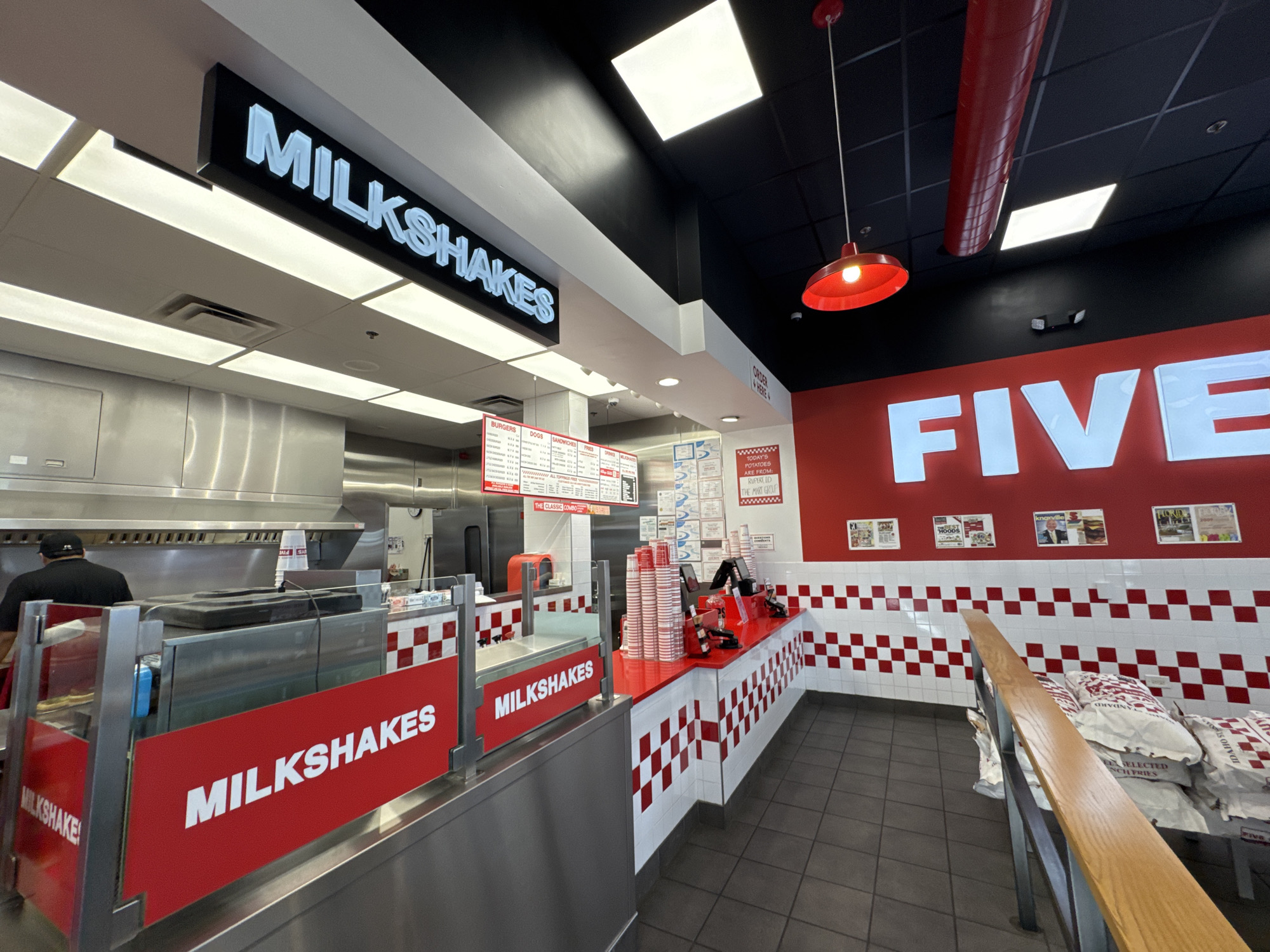 The photo shows the interior of a Five Guys restaurant, likely located in Bloomingdale, United States of America. The restaurant is in the middle of service; one employee is visible behind a stainless steel counter marked "MILKSHAKES." The order counter is lined with red and white checkered tiles, and stacks of cups and ordering equipment are present.
On the wall behind the counter, the word "FIVE" is displayed in large white letters. Numerous documents and notices are posted on the wall. Bags of potatoes are stacked in the seating area, which is marked with a wooden railing, next to the wall.
Overhead, a black ceiling features recessed lighting and a red industrial-style light fixture. Signs are visible above the counter indicating the order placement location.