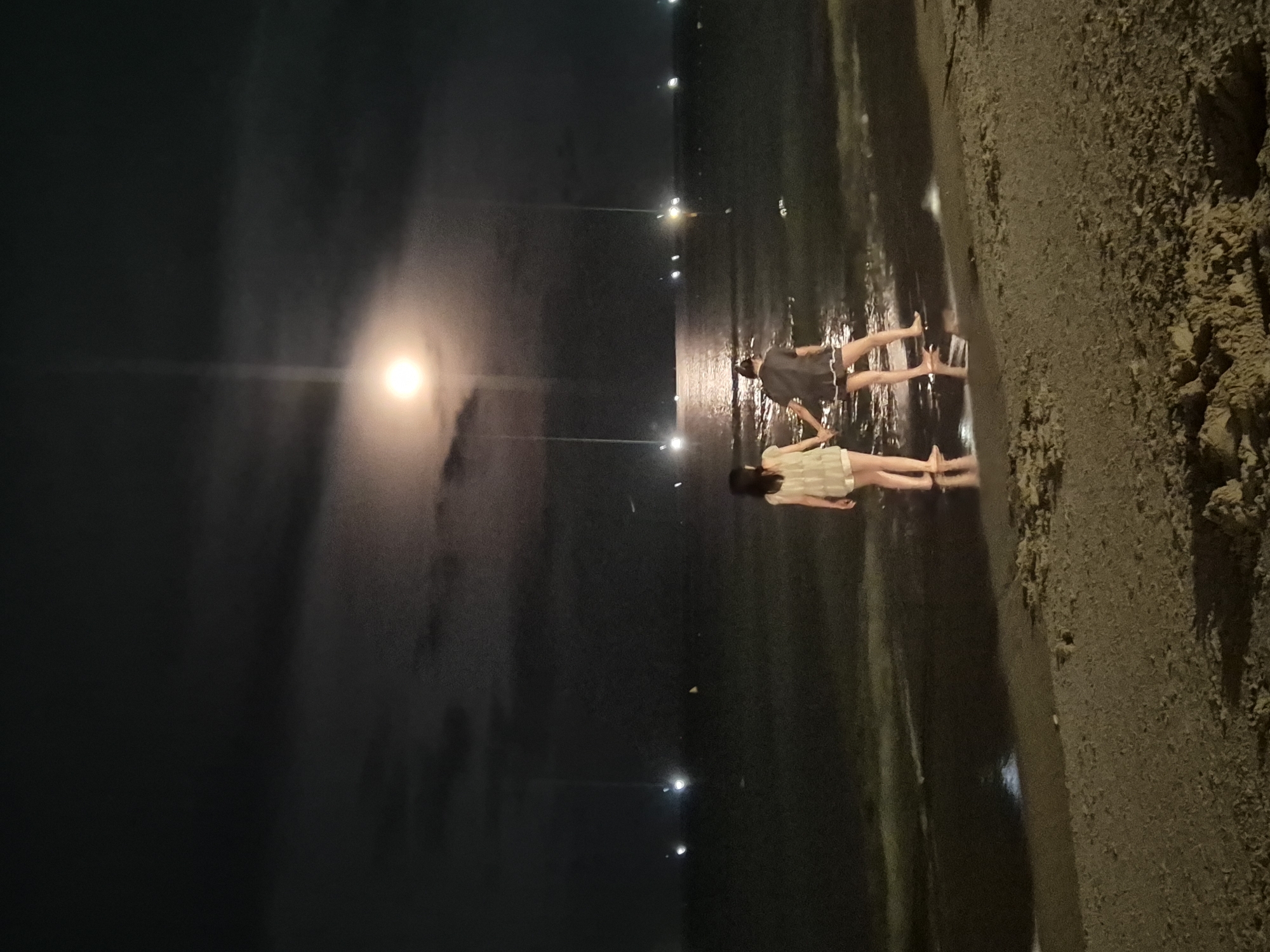 The image depicts a nighttime scene on a beach in Da Nang, Vietnam. Two individuals, appearing to be young women, are standing on the wet sand at the edge of the water, holding hands. The moon is visible in the dark, cloudy sky, casting a bright glow that reflects on the water. Distant lights, possibly from boats or the city, dot the horizon. The sand in the foreground is textured, showing footprints and debris. The overall atmosphere is serene and slightly ethereal due to the moonlight.