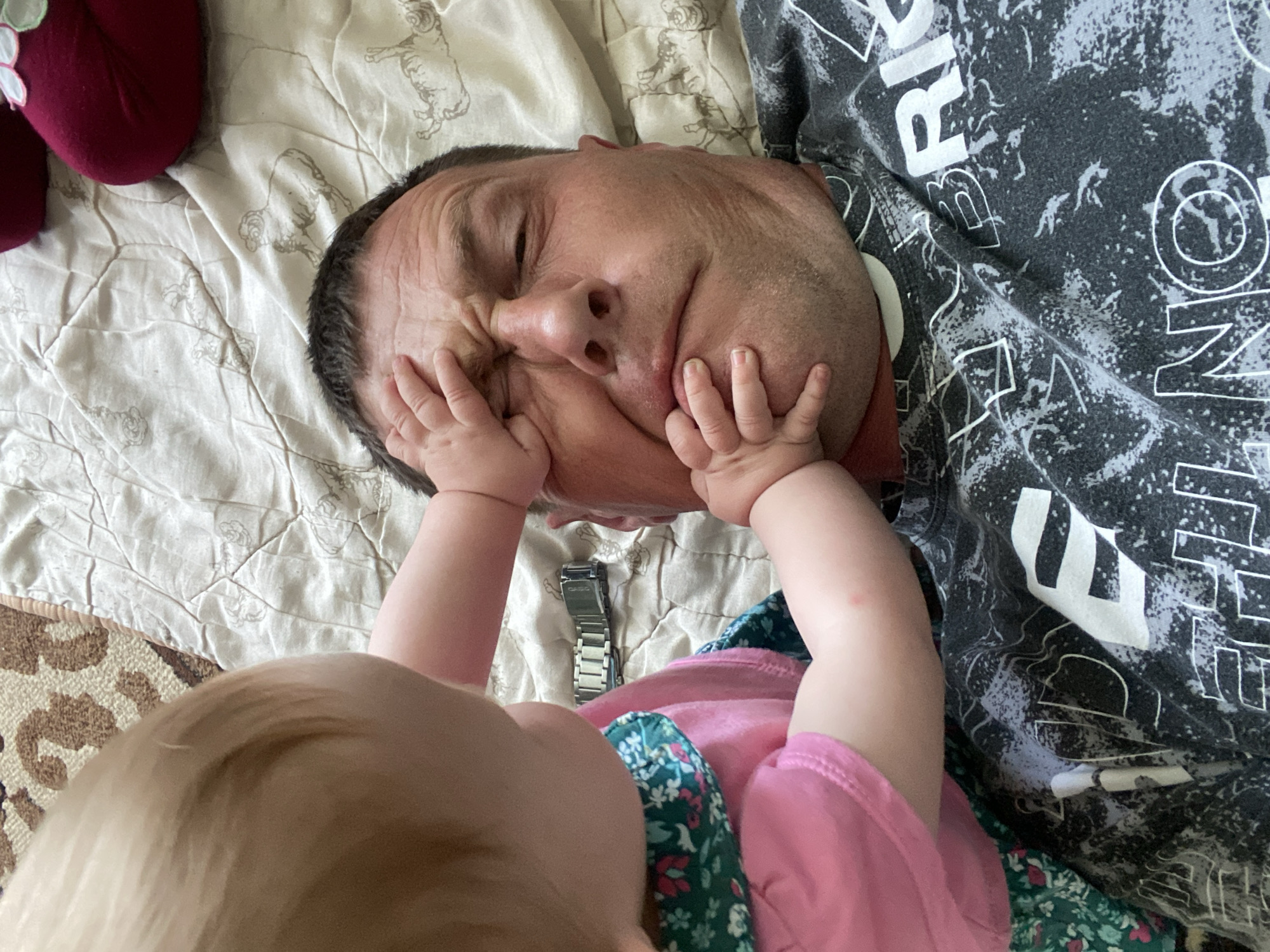 A man lies on his back on a bed with a white patterned sheet, eyes closed, his face being touched by a baby. The baby wears a pink shirt and a floral-patterned dress. The man wears a black patterned shirt. A silver watch lies on the bed. A maroon-colored soft object is in the upper left. The location is not discernible from the image.
