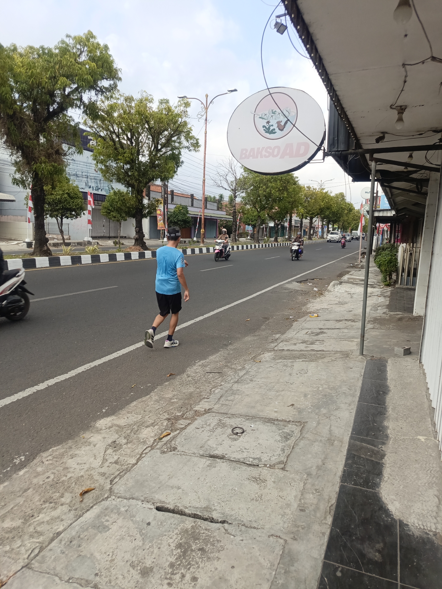 The image captures a street scene in Desa Gunungsimping, Indonesia, with a man walking along the sidewalk next to a road. The man, wearing a blue t-shirt and black shorts, is walking away from the camera towards the right side of the frame. He appears to be casually strolling. The road is paved and has white and black painted lines marking the lanes. Several motorcycles are seen on the road, with some of them in motion. On the left side of the road, there are trees and buildings. One building has a circular sign that reads "BAKSO AD" with an image of a rooster and plants. The sidewalk is made of concrete slabs, with some of them cracked and uneven. The environment suggests a typical day in a town with moderate traffic and pedestrian activity. The weather appears to be overcast or hazy.