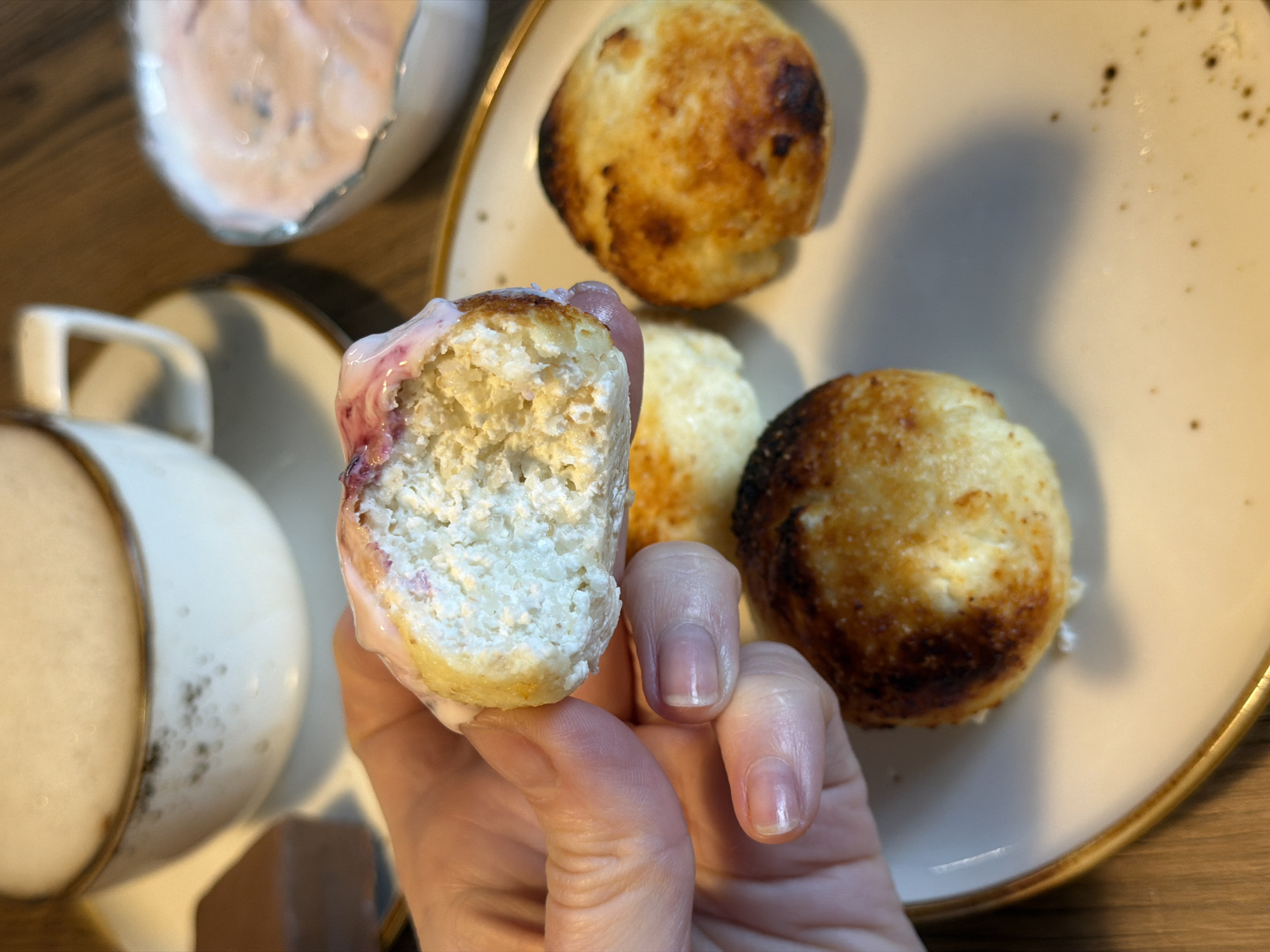 A hand is holding a half-eaten syrniki with pink sauce in a naturally lit environment. In the background, there is a plate with three more syrniki, a small bowl with the same pink sauce, and a cup of coffee. The items are resting on a wooden surface. This image could have been taken in Perm, Russia, based on the popularity of syrniki in Russian cuisine.