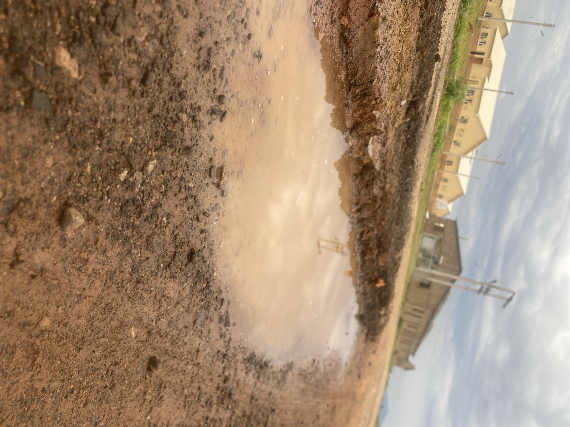 The image depicts a close-up view of a muddy puddle on a dirt road. The puddle is a light brown color and reflects the sky. The surrounding ground is composed of dirt, gravel, and small stones. In the background, there are several buildings with light-colored roofs and walls, possibly residential homes. Utility poles are visible in front of the buildings. The sky is overcast with clouds. The scene suggests it may have recently rained, creating the puddle. The time of day appears to be daytime, given the ambient light. The location is identified as Bulamare, Nigeria. No people are visible in the image.