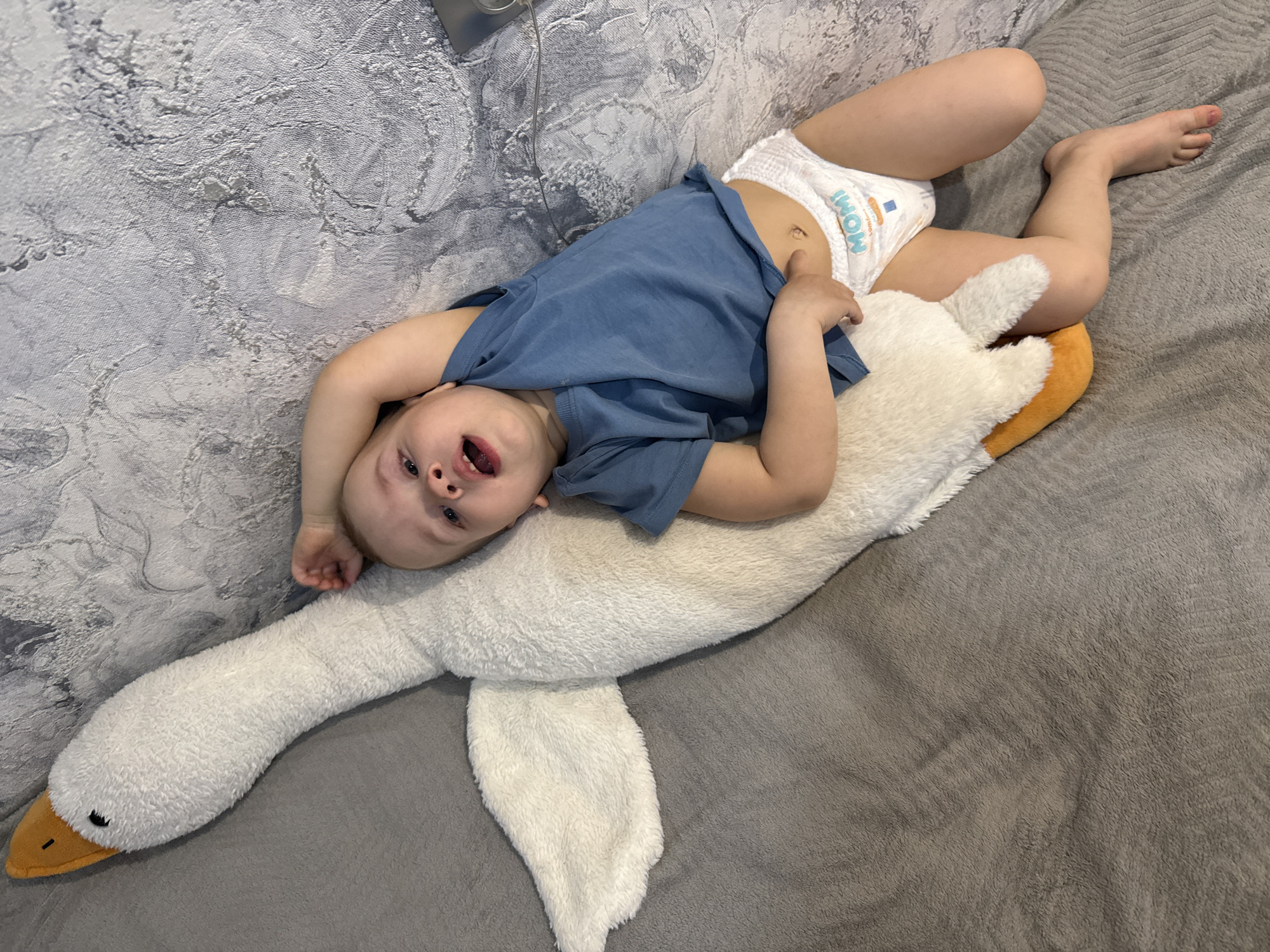A young child lies on their back atop a large, white plush swan. The child wears a blue t-shirt and a diaper. Their head is tilted towards the viewer, and their mouth is open. The plush swan is positioned on a gray blanket, with a textured wall in the background.