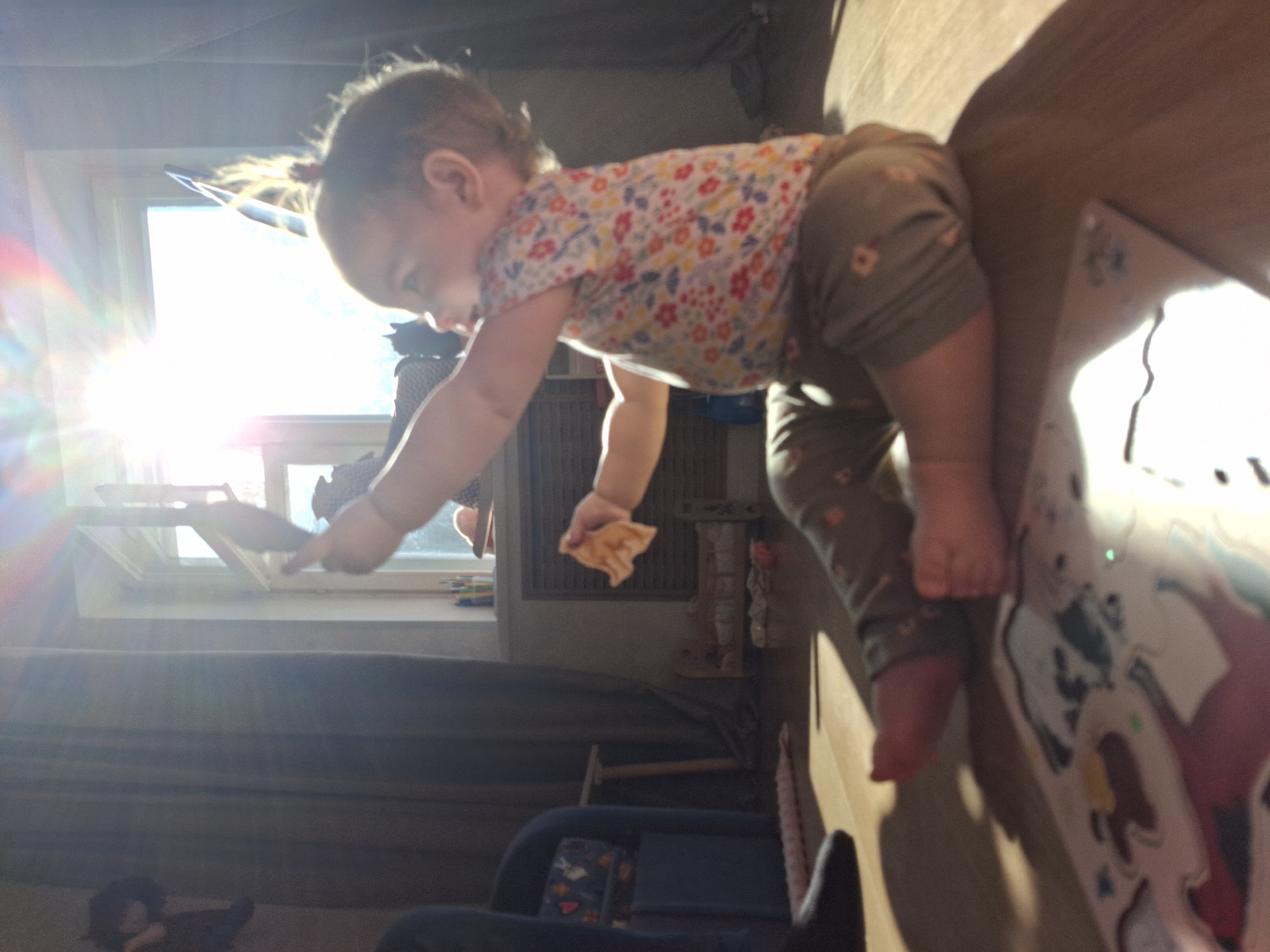 A baby, wearing a floral-patterned top and green pants, is positioned on top of a wooden structure with colorful cartoon illustrations. The baby is leaning forward with one arm extended towards a window where bright sunlight is streaming in. The baby holds a piece of food in its other hand. The location is indoors, likely a residence. A blue chair is visible beneath the window. The city is mentioned in the prompt, therefore this should take place in Chaikovsky, Russia.