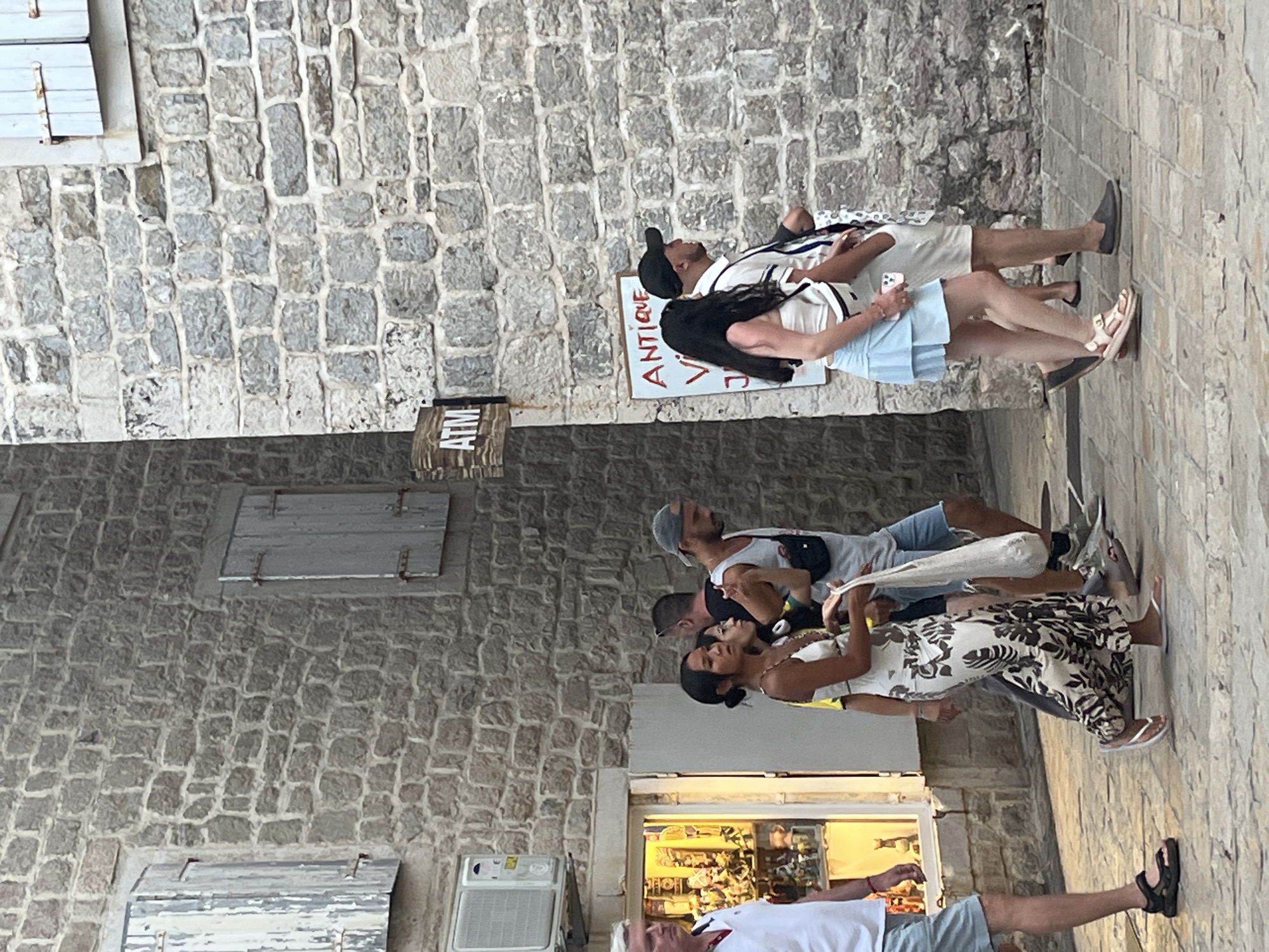 This image depicts a street scene in Budva, Montenegro, likely in the Old Town, characterized by its ancient stone architecture. A group of people are walking along a cobblestone street. On the left, a man in a white polo shirt and blue shorts is walking towards the foreground. Next to him, a woman in a long, black and white patterned dress is walking away from the camera, holding a white bag. Behind her, a man in a grey tank top and blue shorts, and another person, are also walking. Further ahead, a couple is walking away from the camera. The woman is wearing a white top and a light blue skirt, while the man is wearing a light-colored striped shirt and white shorts.
The buildings in the background are constructed from rough, grey stone, with weathered wooden shutters on the windows. A sign with "ATM" is visible on the stone wall. Another sign, likely for a shop, reads "ANTIQUE." An air conditioning unit is mounted on the exterior wall of one building. The lighting suggests it is daytime, possibly late afternoon. The overall atmosphere is that of a bustling tourist area within a historic European town.