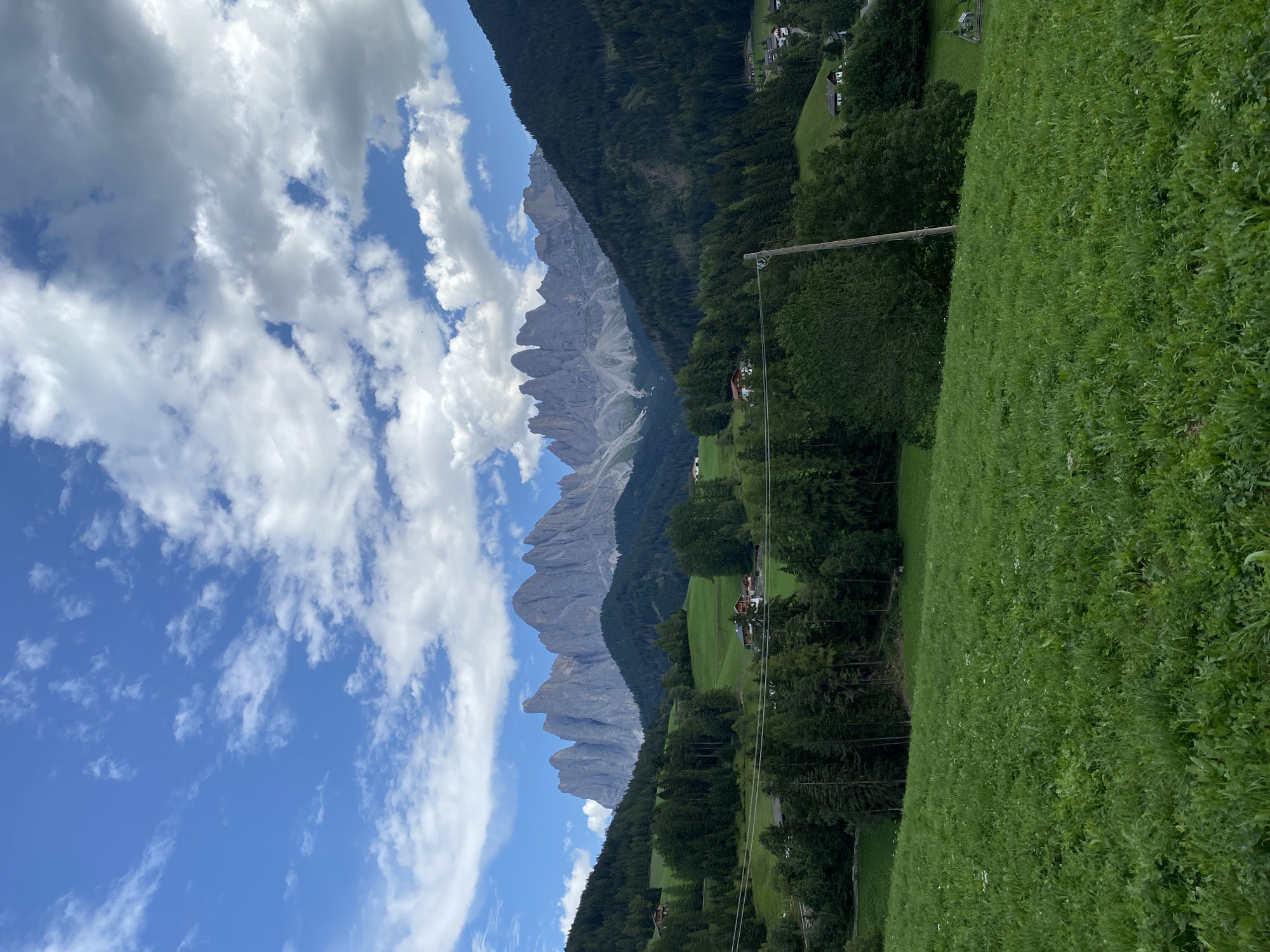 The image depicts a scenic view of St. Magdalena - Santa Maddalena, Italy, under a partly cloudy sky. The foreground is dominated by a lush green meadow with scattered white wildflowers. A steep, tree-covered slope rises from the meadow on the left, transitioning into rolling green hills dotted with a few houses. In the mid-ground, more trees and houses are visible, with a winding road or path visible in the lower left. The background features a dramatic mountain range with jagged peaks, likely the Dolomites. The sky is a vibrant blue, interspersed with fluffy white and gray clouds. A wooden utility pole stands prominently in the middle ground on the right side of the frame. There are no people visible in the image.