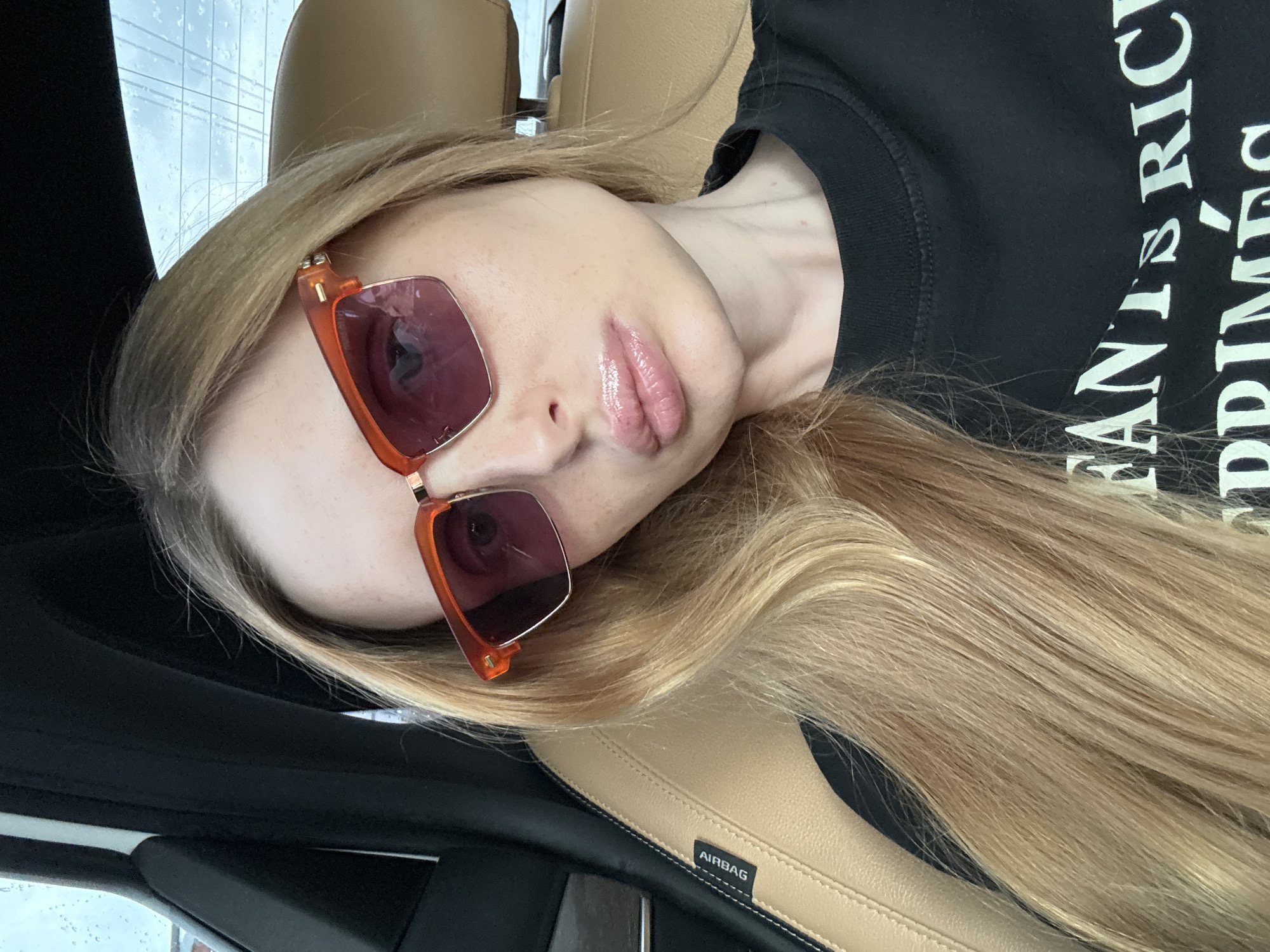 A young woman with long blonde hair is pictured inside a car, wearing orange-rimmed sunglasses and a black t-shirt. The car's interior is visible, with a tan leather seat and a black headrest. A label on the car seat reads "AIRBAG". The background shows a cloudy sky through the car's window. The woman's makeup is prominent, with shiny lipstick and visible freckles.