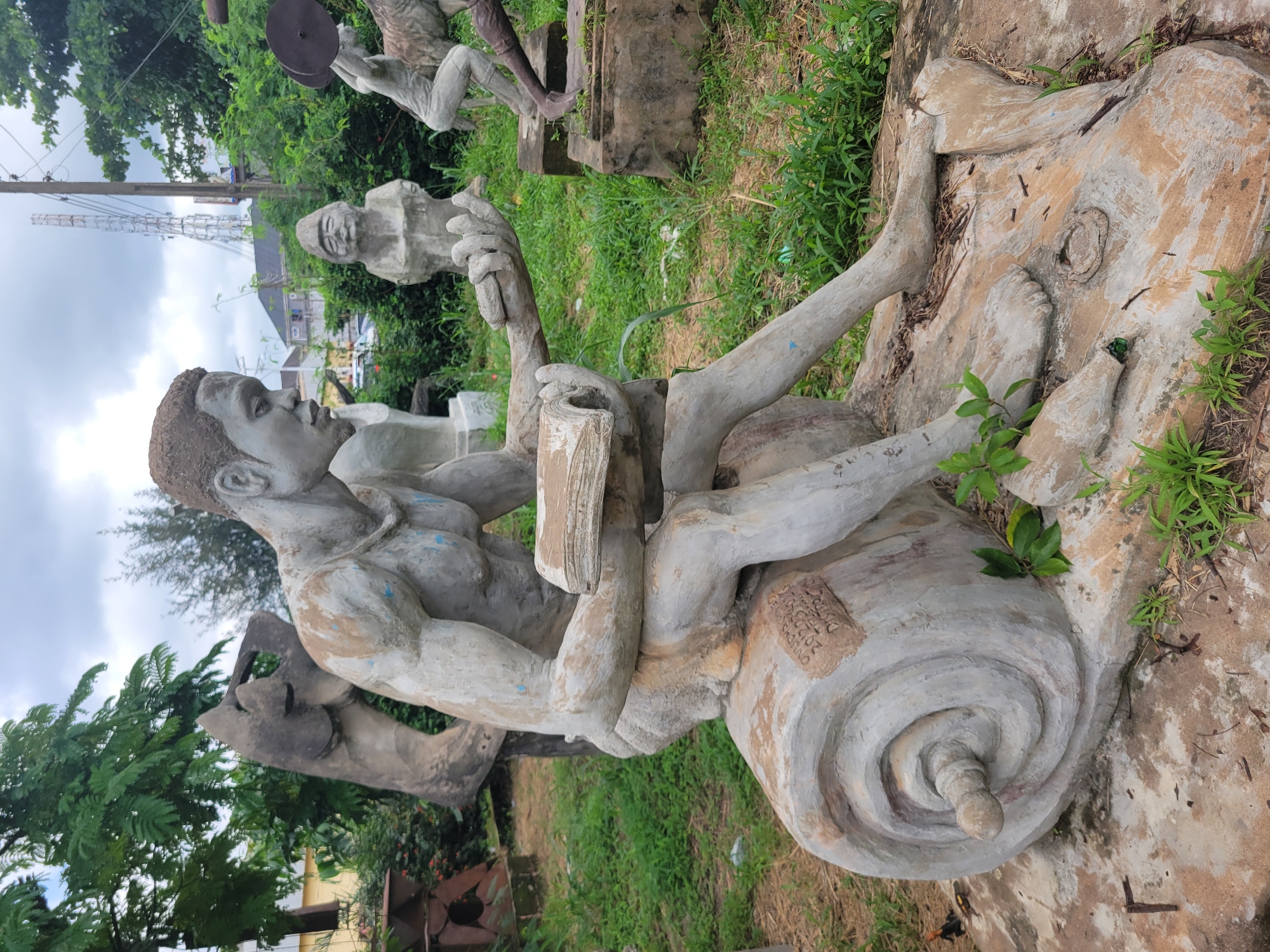 In the city of Egbagu, Nigeria, an outdoor scene displays multiple concrete sculptures amidst overgrown green vegetation and scattered debris. The central and most prominent sculpture is that of a seated male figure, with a weathered, light gray finish showing patches of brown and blue discoloration. The figure has muscular arms and legs, and his head is turned to the side, with his eyes focused downwards. He holds an open book in his lap with both hands. The figure is seated on a large, spiral-shaped base that resembles a scroll, also made of concrete and showing signs of wear.
To the left of the main sculpture, a partial view of another abstract concrete sculpture is visible. In the background, other sculptures are partially obscured by foliage. These include what appears to be a smaller, seated figure with its hands resting on its lap, and a figure in a dynamic pose, possibly lifting weights, to the far right. In the distance, a tall telecommunications tower stands against a cloudy sky, with buildings and trees also visible, suggesting an urban or semi-urban setting. The ground surrounding the sculptures is covered in grass, dirt, and dry leaves, with some visible concrete structures and broken pieces. Two small lizards are present on the ground in the lower left portion of the image.