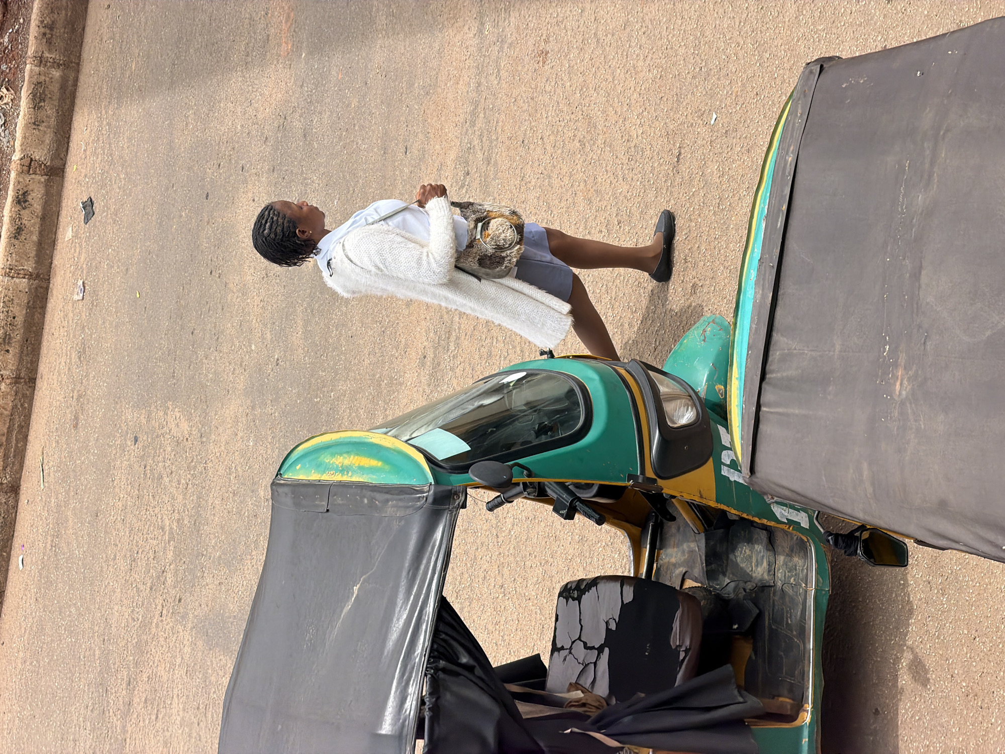 In Bukuru, Nigeria, a woman with braided hair walks next to a green and yellow auto-rickshaw. She is wearing a white fuzzy cardigan over a light blue shirt and a gray skirt, and carries a brown patterned bag. The auto-rickshaw, a common mode of transport, is parked on a dusty, paved road. The vehicle features a black canopy over the passenger seat and a black canopy extending over the front. The road surface is sandy brown, and a curb runs along the top of the frame, with some debris visible on it. The perspective is from an elevated viewpoint looking down.