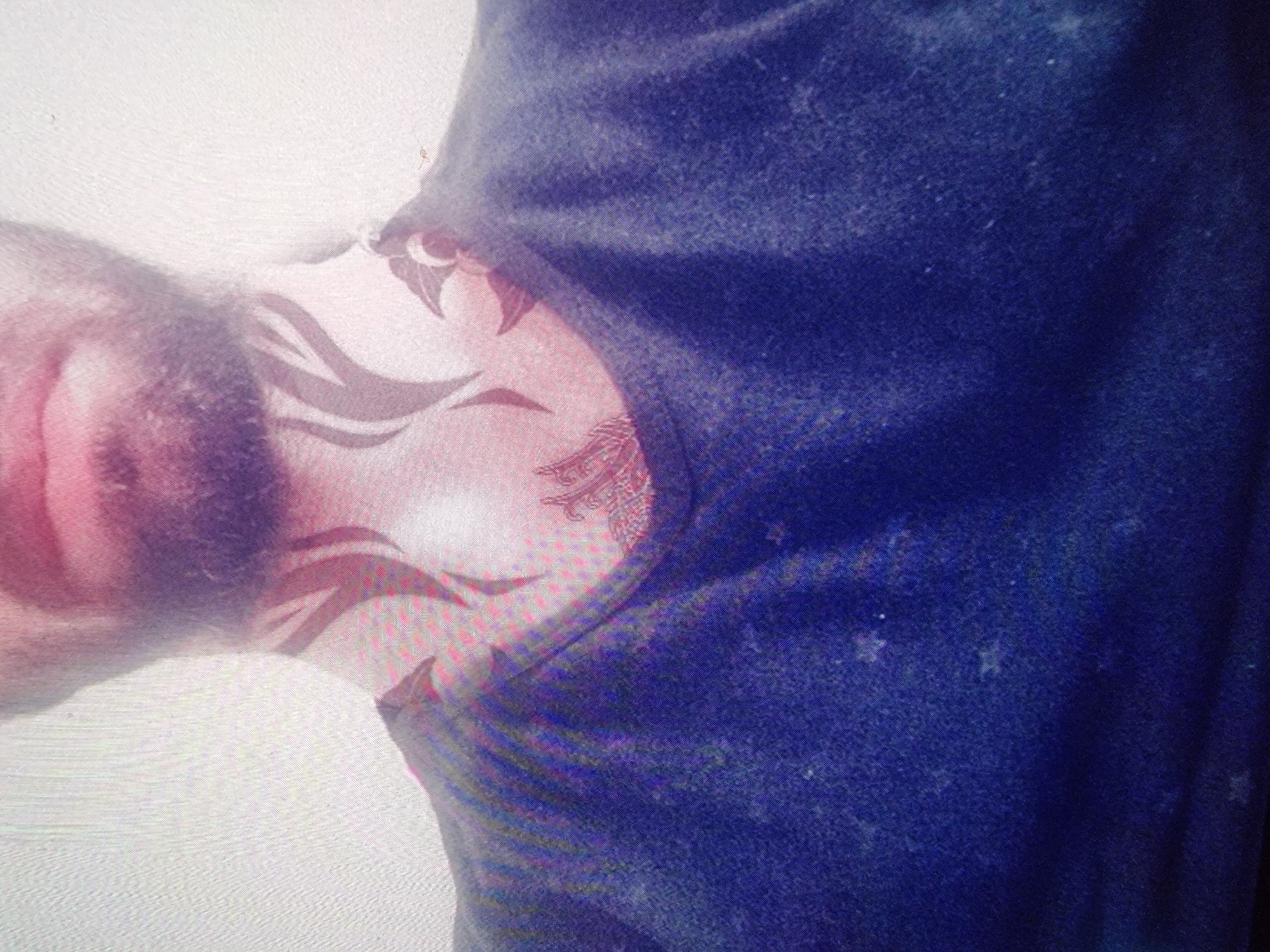 A close-up, slightly skewed photograph shows a person from the chest up, with a dark blue t-shirt. The person has a beard and their neck has a visible tattoo. The background appears to be a plain white wall. The location is Unknown, Unknown.