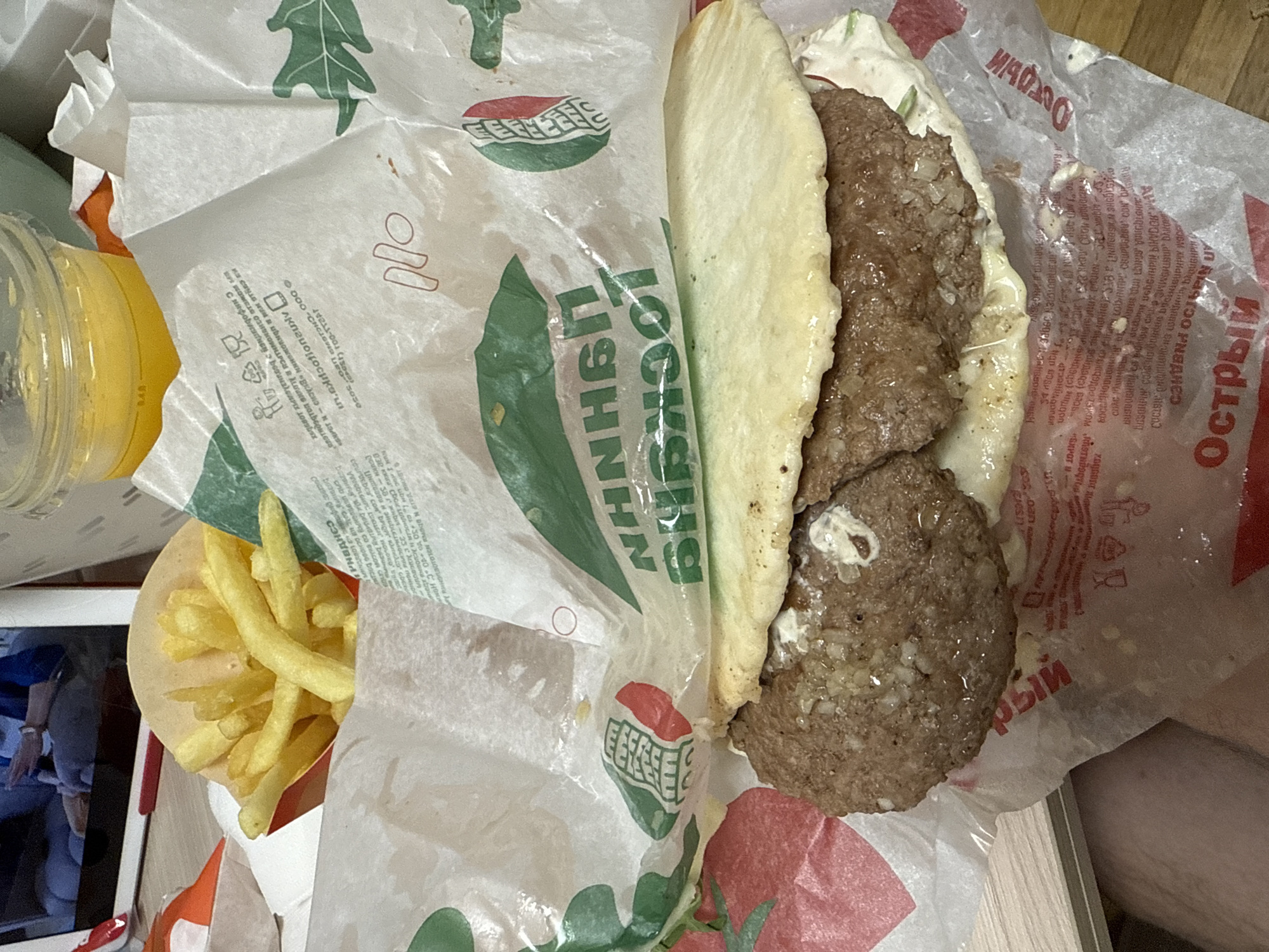 This is a top-down, close-up shot of a fast-food meal. The main subjects are a hamburger and french fries, both unwrapped and placed on branded paper. The hamburger is open, revealing two patties, a white sauce, and some chopped onions. The french fries are golden brown and appear to be a side dish. The background is dominated by the red and white branded paper wrappers from the fast-food establishment. Some text is visible on the wrappers, including the words "Острый" (Spicy) and "Цитвата" (Citrus). The lighting suggests it might be daytime, but no specific time or weather cues are apparent. The overall scene is casual and appears to be in a relaxed setting, likely a home or a picnic, given the informal presentation of the food. No people are visible in the frame, but a partial view of a person's leg is present at the bottom right. There is also a yellow cup with a beverage, likely juice, on the left side of the image. The image has a slightly grainy texture, suggesting it was taken with a mobile phone.