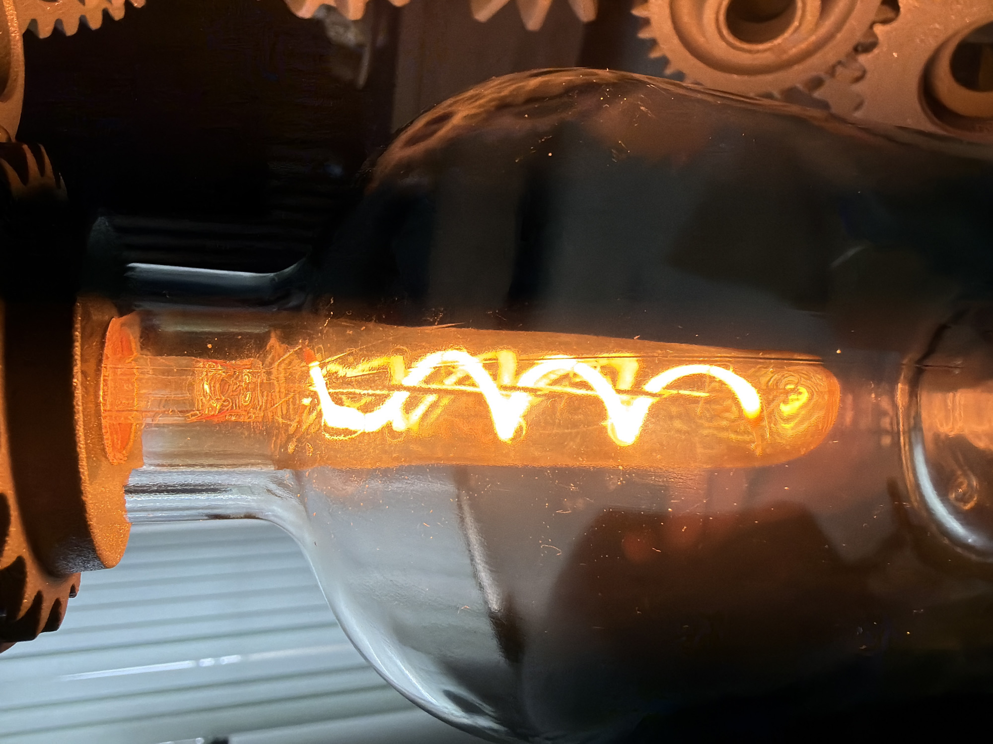 A close-up, horizontally oriented photograph captures an illuminated Edison-style light bulb, showcasing its glowing, intricate coiled filament. The filament emits a bright, warm orange-yellow light, illuminating the clear glass bulb and casting a soft glow on its surroundings.
The light bulb appears to be part of a larger fixture with industrial or steampunk aesthetics. Partially visible in the upper and left portions of the image are sections of metallic gears with ridged teeth, suggesting a mechanical setup. These gears are dark grey or bronze in color, contrasting with the bright light. Reflections of the glowing filament are visible on the curved glass surface of the bulb, creating patterns of light and shadow.
The background is predominantly dark, emphasizing the brightness of the bulb. In the lower left quadrant of the main image, and more clearly visible in one of the hint crops, there is a light-colored, horizontally ribbed surface, possibly a shutter, blind, or wall panel, contrasting with the darker elements. The overall setting implies low-light conditions, with the bulb acting as the primary light source. There is no visible text or discernible activity beyond the bulb being lit. The image evokes a sense of nostalgia or vintage industrial design.