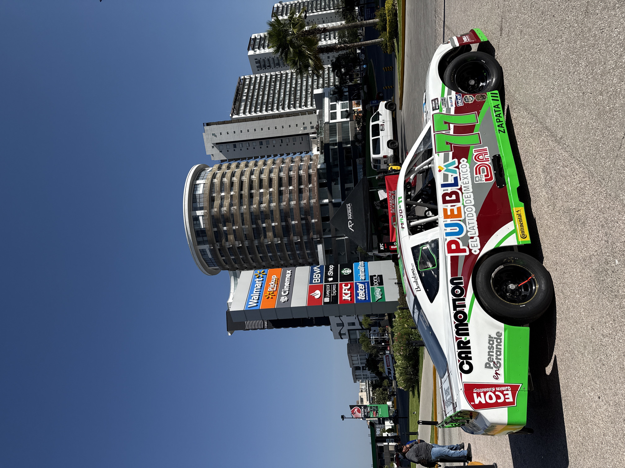 The image shows a race car parked on an asphalt surface, likely a parking lot or street. The car has a white base color with green and maroon accents and various sponsor logos like "CAR MOTION," "ECOM," "PUEBLA," "Continental," and "telcel." The number 77 is prominently displayed on the car. In the background, there is a multi-story cylindrical building with a unique architectural design and several taller, more standard apartment buildings. A sign in the background shows logos for a shopping center or plaza, with logos for Walmart, Cinemex, BBVA, KFC, and other brands. A white van is also visible between the buildings. In the lower left, a man is lying down on a bench or a platform. The sky is a clear, bright blue. The overall impression is that of a race car on display near a commercial area in San Bernardino Tlaxcalancingo, Mexico.