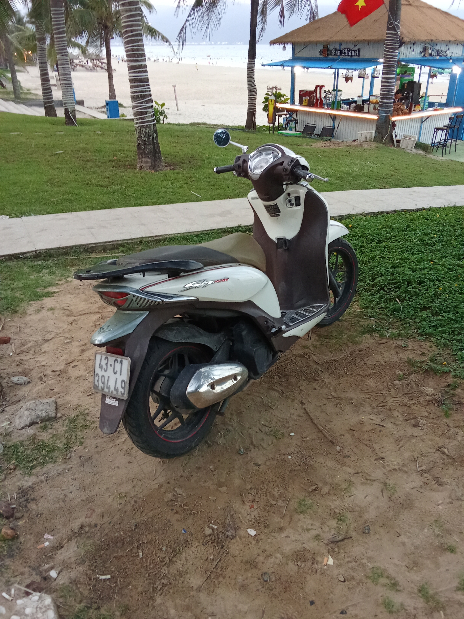 A white and brown scooter is parked on a sandy area next to a grassy patch with a concrete walkway. In the background, there is a beach with people and a bar-style building with a thatched roof. Palm trees are visible on either side of the building. The scooter has a license plate that reads "43-C1 394.49". The image appears to have been taken in the city of Đà Nẵng, Vietnam, given the presence of palm trees, beach, and a bar reminiscent of beachside establishments often found in the region.