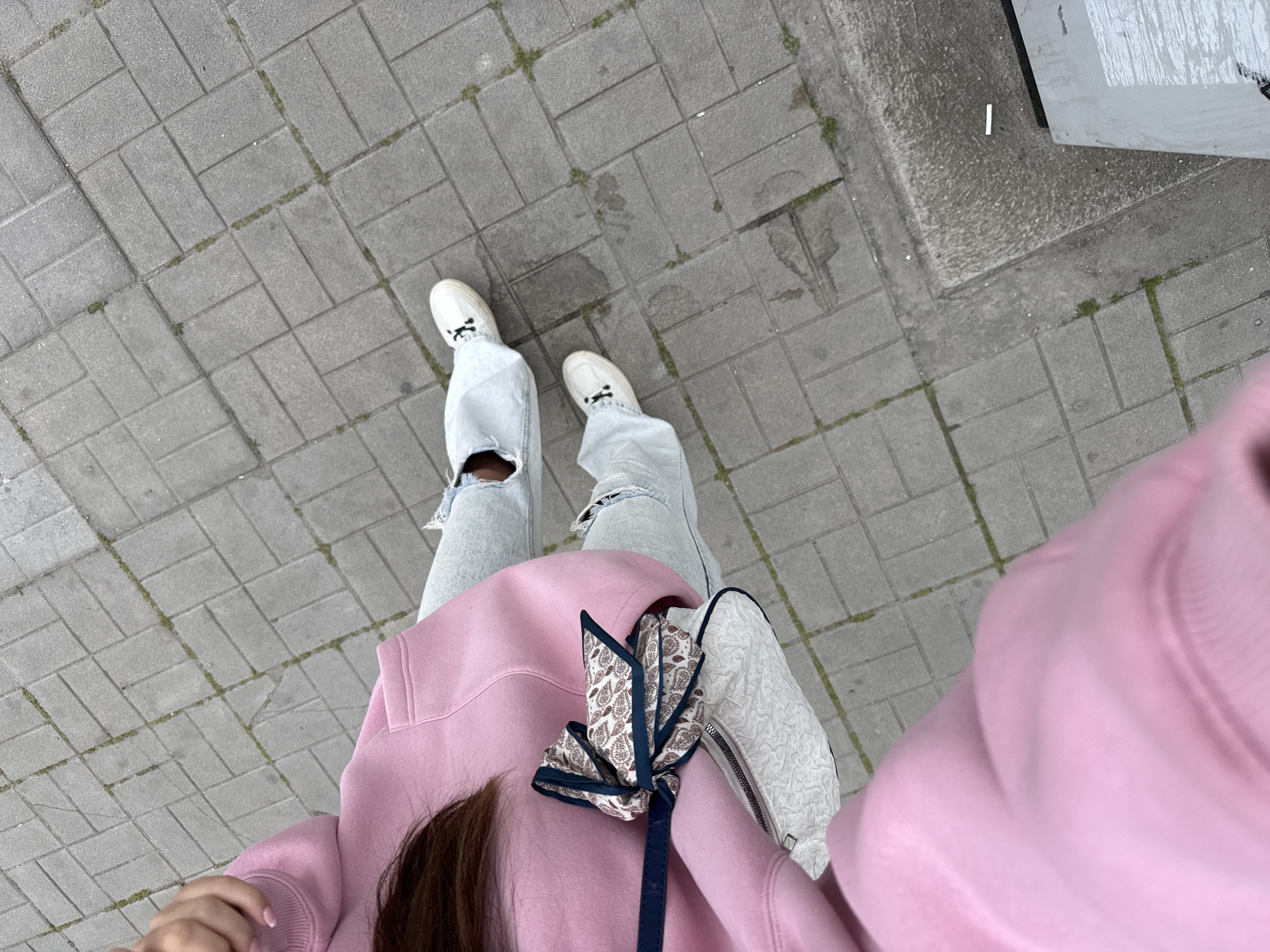 The image depicts a person standing on a grey brick-paved surface. The person is wearing white shoes, light blue ripped jeans, and a pink hoodie. A white handbag with a floral patterned scarf tied to it is slung across their shoulder. The background includes a concrete curb. The image appears to have been taken from a first-person perspective. The location appears to be a pedestrian walkway in an urban area. Without any additional information about landmarks or context visible in the image, it's impossible to conclusively establish the precise location within Plovdiv.