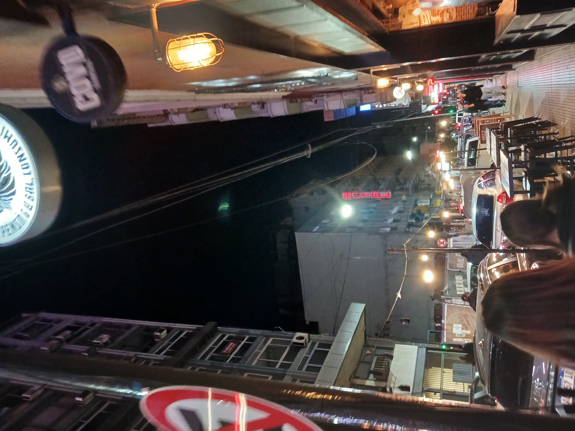 The image shows a street scene at night in Buenos Aires, Argentina. The buildings on the left side of the image are dark, with some lights visible. A red neon sign with the words "HOTEL" is visible on a building in the distance. On the right side of the image, there is a street with several cars parked and people walking on the sidewalk. Tables and chairs, possibly from a restaurant, are also visible on the sidewalk. The presence of Argentine license plates on cars suggests the location. Streetlights illuminate the scene, creating a mix of shadows and light.