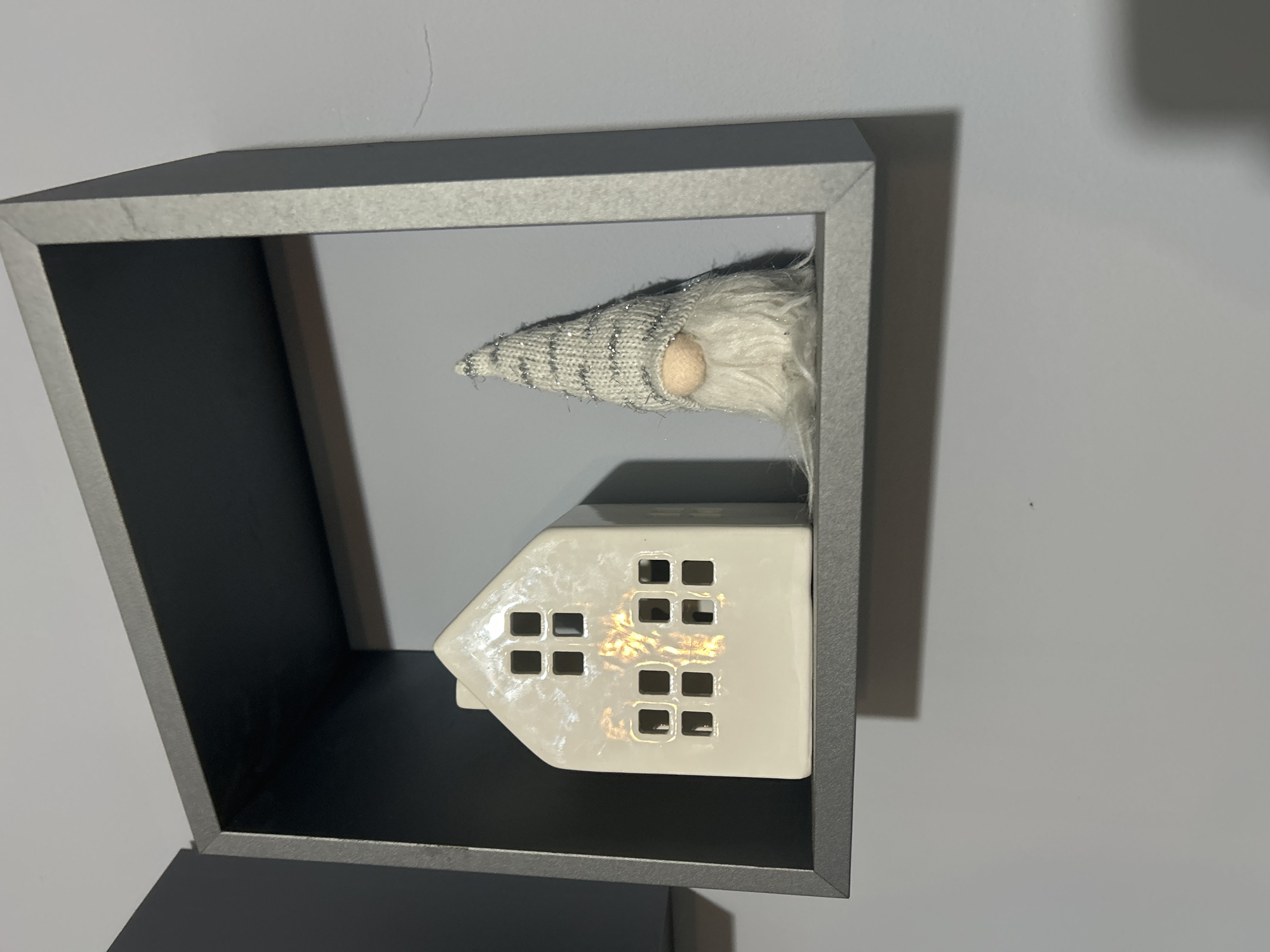 A close-up shot depicts a gray cube shelf containing a white ceramic house with window cutouts and a gnome with a gray and white striped hat. The house sits on the lower portion of the shelf, and the gnome's hat extends upward. The shelf is mounted on a light gray wall. The location is Unknown, Unknown.