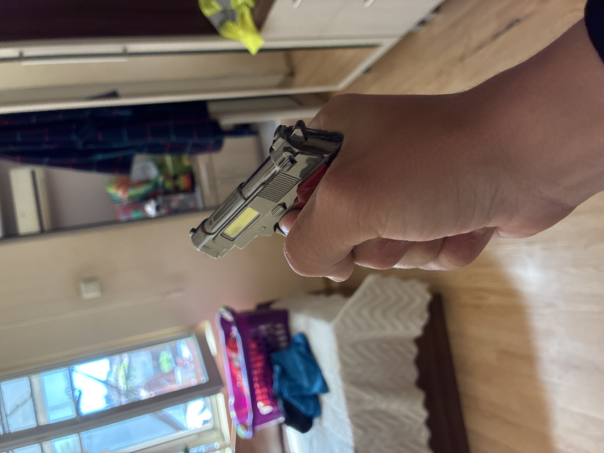 A human hand, primarily the palm and fingers, is centrally positioned, holding a small, metallic replica handgun. The replica strongly resembles a Beretta M9 or 92FS, featuring detailed rear sights, a hammer, and a textured slide. The hand holds the item firmly, pointing it slightly upward and away from the viewer.
The setting is an indoor domestic environment, likely a bedroom or living area. In the background, to the left, is a wardrobe or dresser with an open section revealing blurred dark plaid fabric and other items. A bright yellow object is partially visible on top of this furniture. To the right, part of a bed with a white bedspread featuring a distinct zig-zag pattern is visible.
In the bottom left of the frame, a window shows bright daylight and reflections of green foliage and trees outside, indicating it is daytime. Near the bed, a purple basket and a blue piece of cloth or garment are discernible. The flooring throughout the visible area is light-colored wood. A shadow, likely cast by the hand and gun or an adjacent object, extends across the wooden floor in the bottom right corner. No specific activity beyond holding the item is depicted, and no emotions can be inferred. There is no visible text in the image.