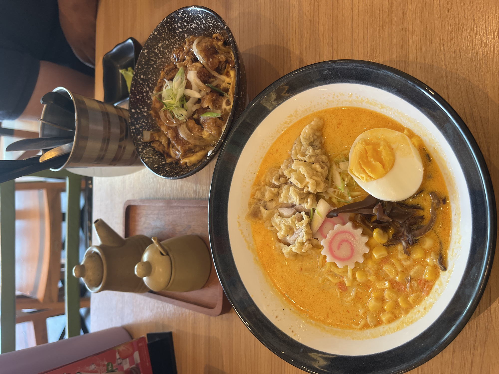 A high-angle shot depicts a table setting with two bowls of food in what appears to be a restaurant in Lhokseumawe, Indonesia. On the right is a bowl of ramen with a yellow broth, topped with corn, a sliced hard-boiled egg, seaweed, meat (likely chicken), and a narutomaki (fish cake). Next to it, on the left, is another bowl containing a rice bowl (Katsudon). A metal container with utensils, a small black dish, and two sauce containers on a wooden tray are also visible on the table. The table itself is light-colored wood. In the background are chairs, suggesting an indoor dining area. A person is partially visible on the top left of the frame.