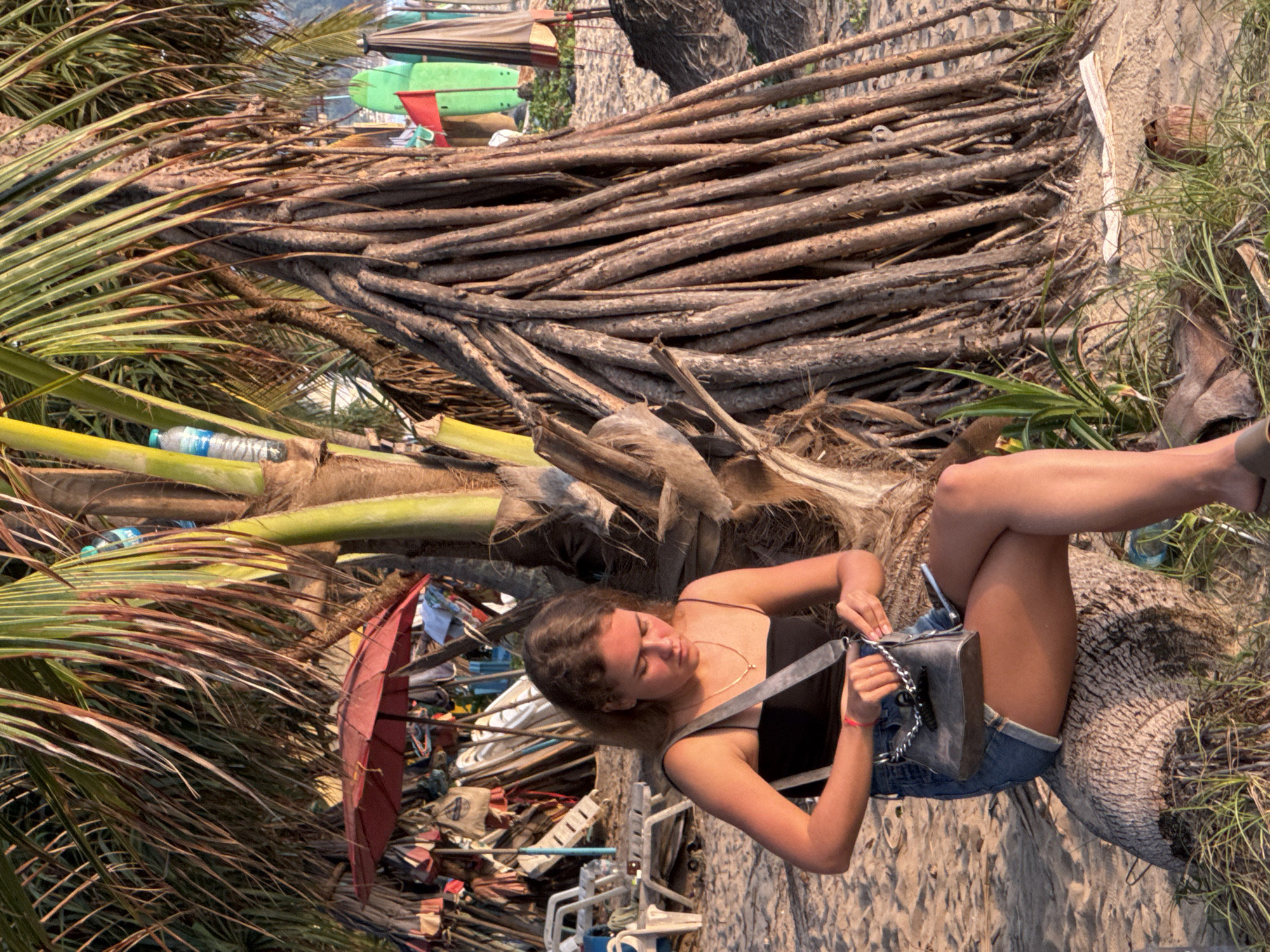 A young Caucasian woman with blonde hair is sitting on a log on a sandy beach in Karon, Thailand. She is wearing a black spaghetti strap top and denim shorts. She has a small grey purse with a chain strap in her hands, and appears to be looking at something inside it. Behind her, a palm tree with exposed roots is prominent, with more palm fronds and trunks creating a backdrop. In the background, there are beach chairs, umbrellas, and surfboards, suggesting a tourist beach setting. The lighting indicates it is late afternoon or early evening.