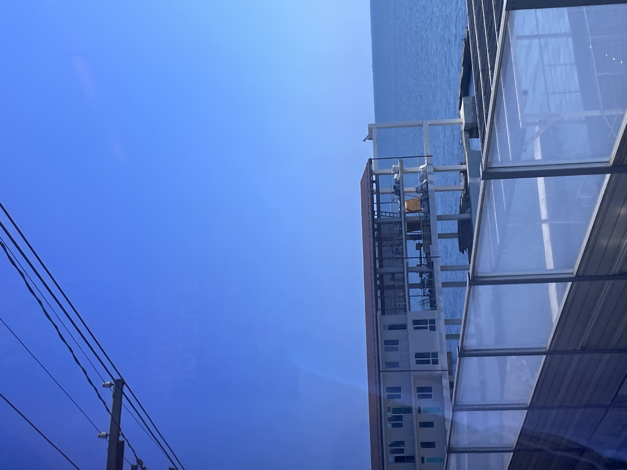 This image captures a waterfront building along the Black Sea coast in Sochi, Russia. The scene is dominated by a clear, vibrant blue sky. In the foreground, a modern structure with glass railings and a slatted roof is visible, suggesting a viewing deck or an elevated walkway. Beyond this, a multi-story building stands on stilts over the water. This building features a light-colored facade with numerous windows, and its upper levels are partially enclosed by glass walls, revealing tables and chairs, indicating it's likely a restaurant or hotel. The water is a deep blue, reflecting the sky. Overhead, power lines crisscross the frame, adding a utilitarian element to the otherwise scenic view. The image appears to be taken from a moving vehicle, possibly a train or bus, given the slight blur and reflection on the glass in the foreground. There are no people clearly visible in the frame.