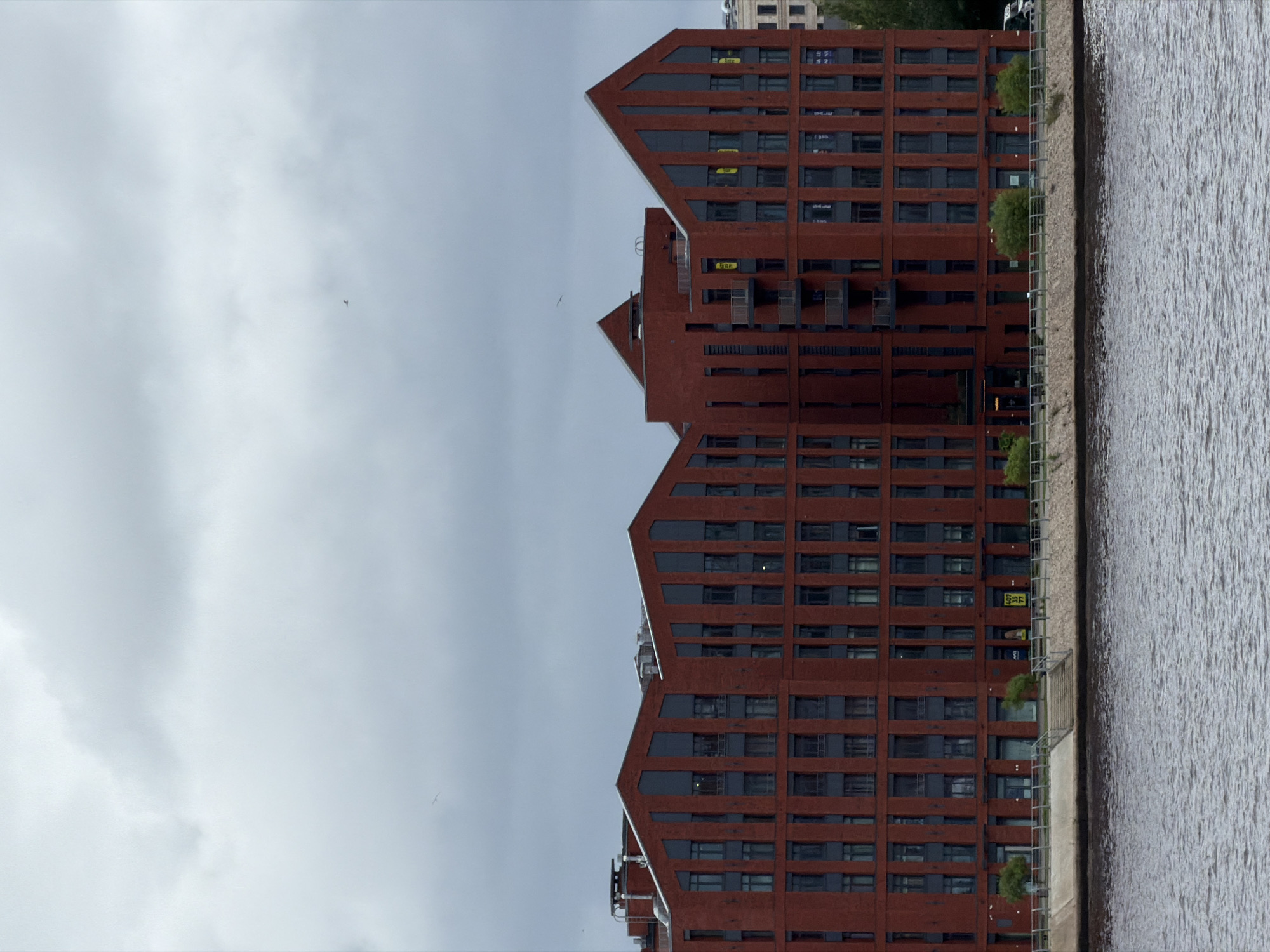 The image depicts a modern, brick-clad apartment building situated on the waterfront in Saint Petersburg, Russia. The building features a distinctive, angular roofline with prominent dark grey vertical accents that frame windows and create a segmented facade. Numerous windows are visible across multiple floors, interspersed with balconies on some levels. The building's reddish-brown brickwork contrasts with the dark grey architectural elements.
Along the waterfront, a paved promenade is lined with a metal fence and dotted with small trees. Stairs lead down to the water's edge, where a body of water, likely a river or canal, occupies the foreground. The water is choppy, indicating a breeze. The sky above is overcast with heavy grey clouds, suggesting a diffused light condition. A single bird is visible in the sky, soaring above the building. Other buildings, appearing more traditional in style, are partially visible in the background to the right, hinting at the urban environment of Saint Petersburg. Signs are visible on some windows of the apartment building, potentially indicating apartments for sale or rent. The overall impression is one of a contemporary residential development integrated into the city's historic landscape.