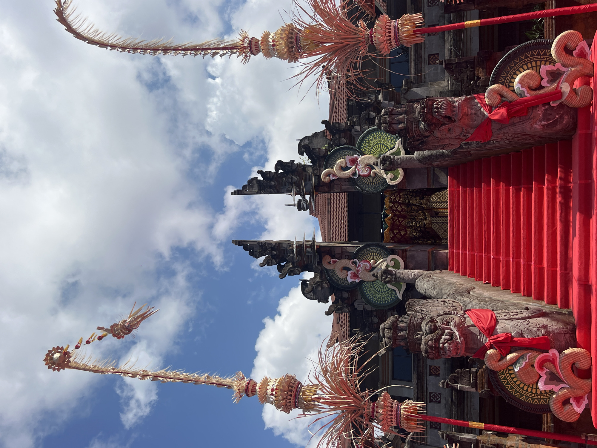 This is a low-angle, full shot of the entrance to a Balinese temple in Denpasar, Indonesia. The temple facade is elaborate, with intricate stone carvings, including two menacing dragon statues flanking a bright red carpeted staircase. The staircase leads up to a grand entrance, flanked by two tall, decorative poles adorned with woven palm fronds and small figures. The sky above is a vibrant blue with scattered white clouds. The scene is festive and appears to be set up for a ceremony or celebration, with the red carpet and decorative elements suggesting a special occasion. The overall impression is one of cultural richness and spiritual significance.