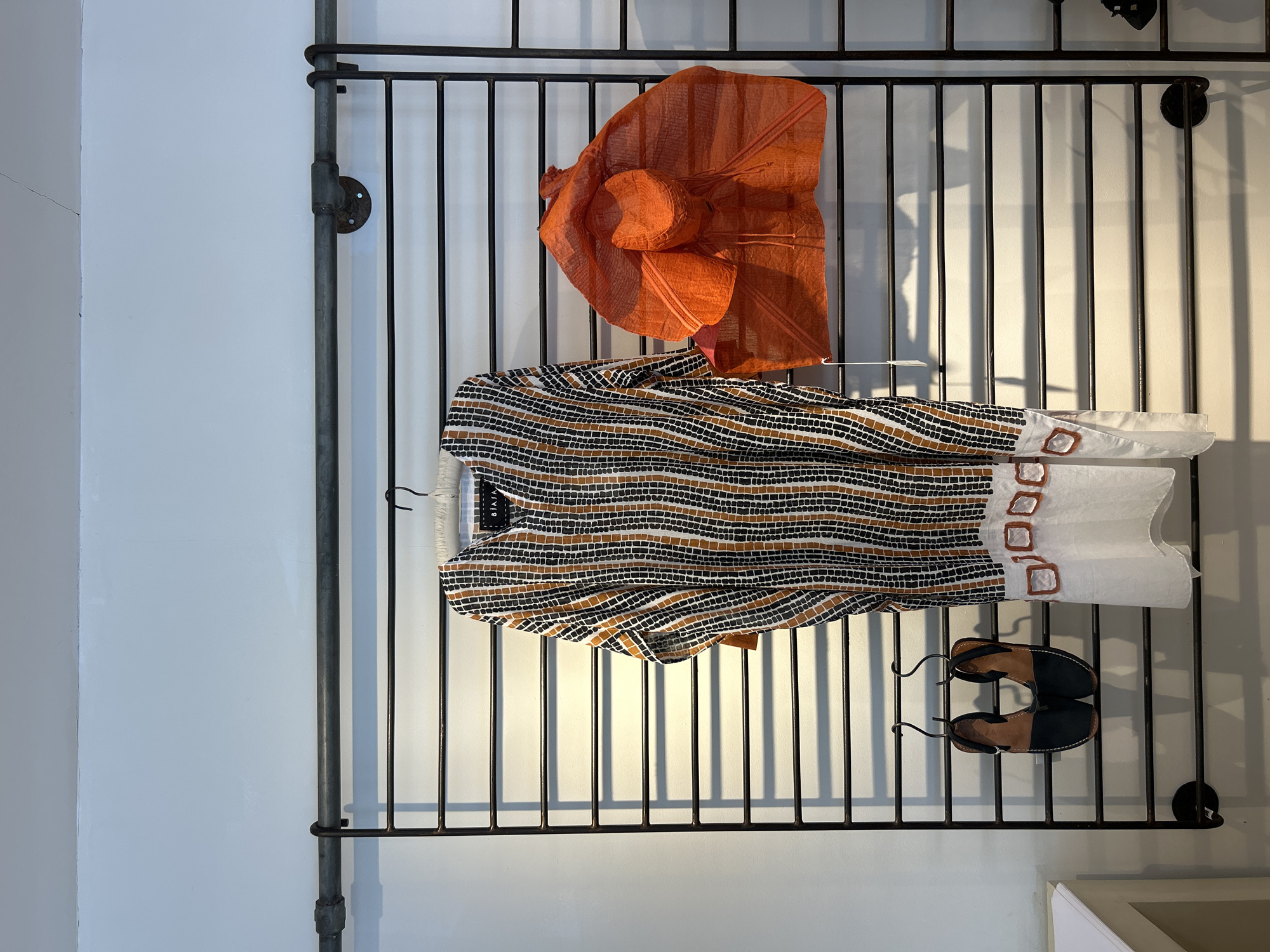 A flat lay of women's clothing and shoes is displayed on a black metal rack against a white wall. The subjects include a folded orange raffia hat and a black, white, and tan striped tunic. The tunic has an asymmetrical hemline with white fabric accents and small square buckle details. A pair of black and tan leather sandals with ankle straps are hanging on hooks below the tunic. The lighting suggests daytime, with shadows cast by the rack onto the wall. The scene appears to be a retail or display setting, possibly in a boutique, given the way the items are presented. There is no discernible text visible in the image. The location context is North Kuta, Indonesia.