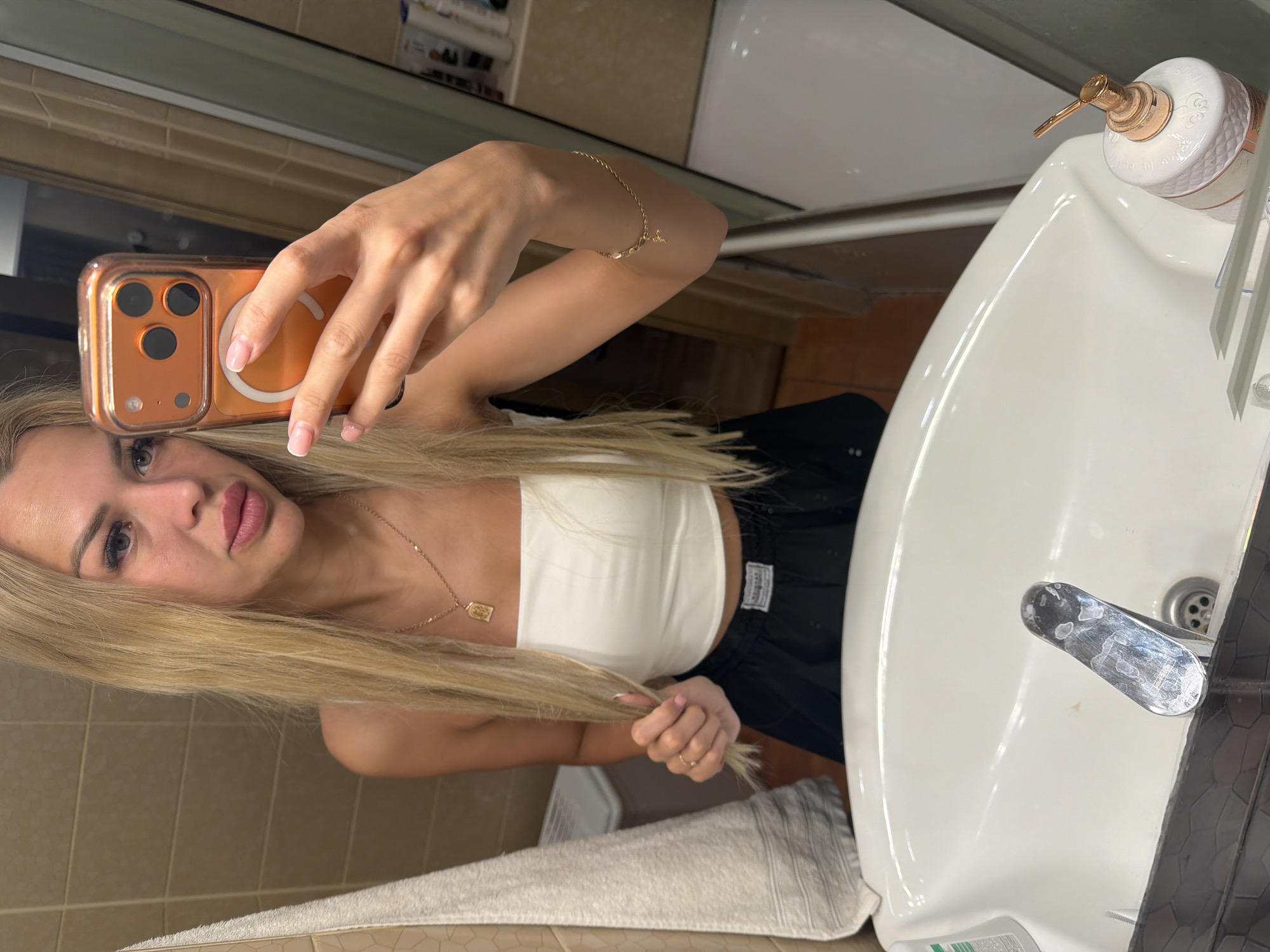 A young woman with long blonde hair, wearing a white tube top and black shorts, takes a selfie in a bathroom mirror. She is holding her phone with her right hand, and her left hand is touching her hair. A gold necklace with a pendant is visible around her neck, and a gold bracelet is on her right wrist. The bathroom features tiled walls and a white sink with a chrome faucet. A white towel hangs to the left of the sink. Bottles of toiletries are visible on a shelf in the background, and a white tub is partially visible to the right. The scene appears to be in an indoor residential setting, likely a bathroom.