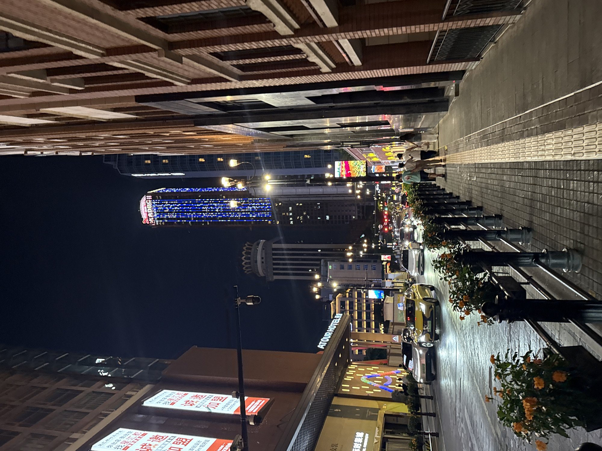 The image shows a nighttime street scene in Yuzhong District, China. The street is lined with tall buildings, some of which are illuminated with colorful lights. Cars are driving on the road, and pedestrians are walking on the sidewalk. There are also decorative elements such as potted plants and streetlights. The overall impression is that of a vibrant and bustling urban environment.