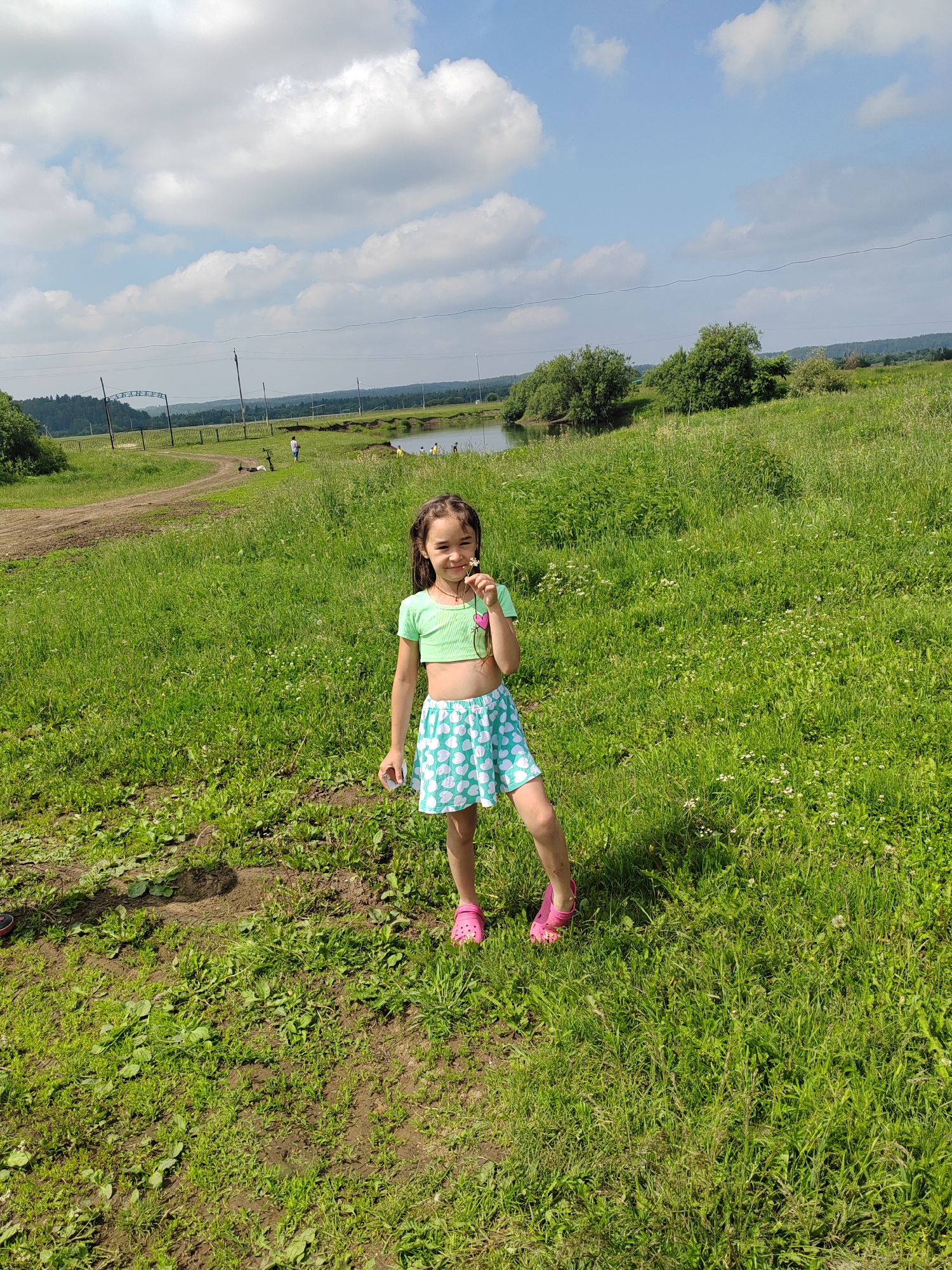In a grassy field under a partially cloudy sky, a young girl stands facing the camera. She has dark hair and is wearing a light green crop top and a light blue skirt with a white heart pattern. She is holding a small white flower and a pink heart-shaped object in her right hand. Her feet are clad in pink Crocs. The background features a dirt path, a body of water, and trees. In the distance, there are faint figures of people and a structure with an archway. The environment appears to be rural.