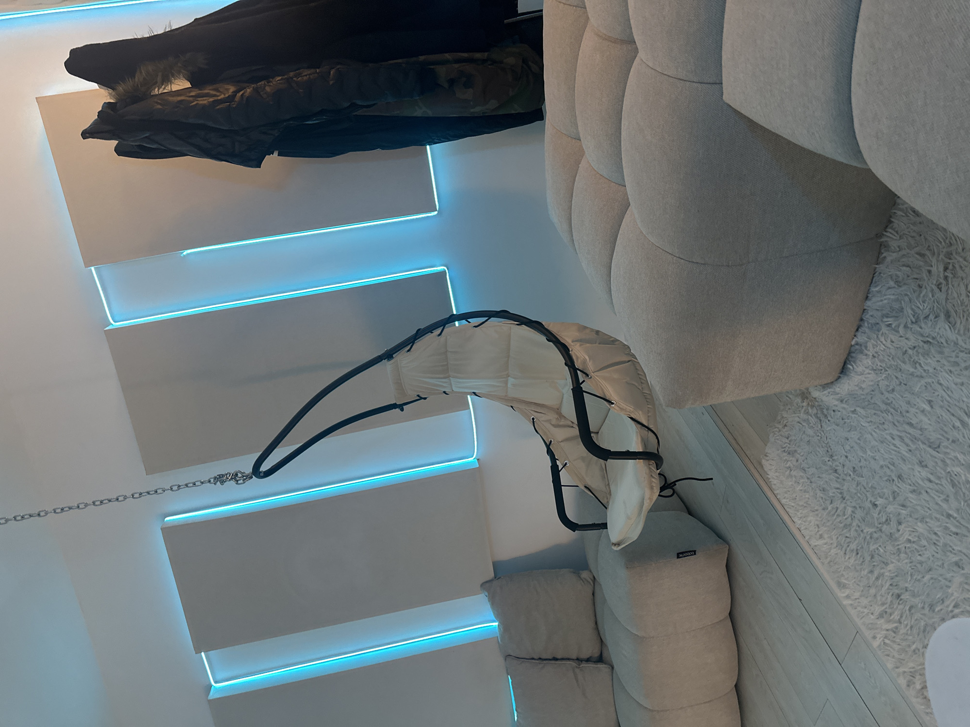 This indoor shot, taken in what appears to be a residential space, features several distinct elements. On the left side of the frame, there is a wall adorned with horizontally stacked panels, each separated by a thin strip of blue LED lighting. A coat or jacket is draped over the top panel.
A hanging baby swing is suspended from the ceiling, occupying the central area of the image. The swing's frame is black, with a white fabric seat and backing. A chain extends from the swing's suspension point, connecting it to the ceiling above.
On the right side of the frame, there is a section of a light gray, tufted sofa, which is partially cropped. Beneath it, there are some stacked cube style pillows or seats on a light colored floor.
The image was taken indoors in Saint-Cloud, France.