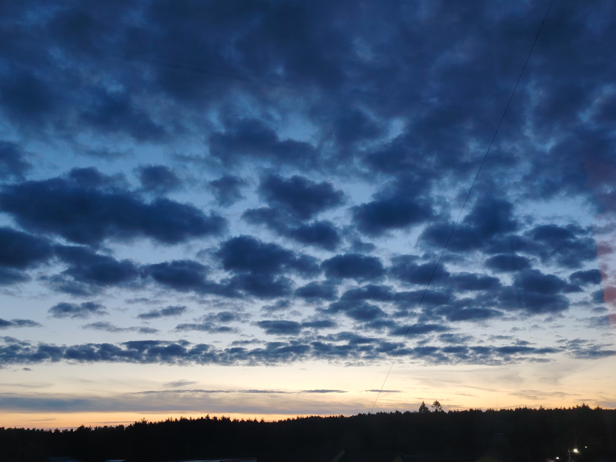 A wide shot captures a dark, cloud-filled sky over a silhouetted treeline in Konakovo, Russia. The clouds are a deep, murky blue with some lighter gray patches. The sky transitions to a lighter blue near the horizon, where a faint yellow and orange glow suggests a setting sun. A thin, dark line, likely a power line or wire, stretches diagonally across the upper right portion of the frame. The treeline below is a solid, dark mass, obscuring any details of the ground or any structures that might be present. The overall impression is one of an overcast evening.