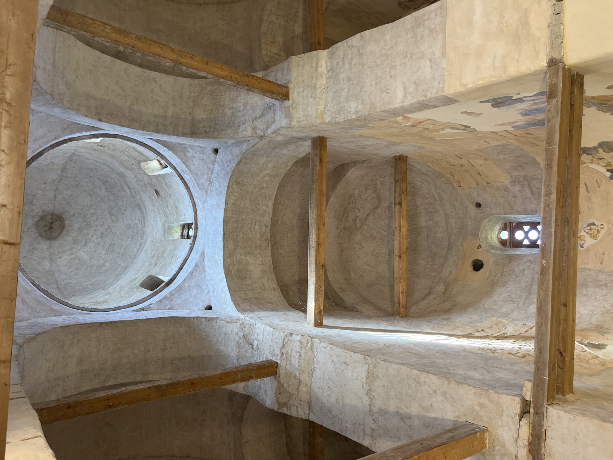The image shows the interior of a domed structure, likely a church, with visible frescoes on the walls and wooden beams supporting the ceiling. The architecture features rounded arches and a central dome with an opening at the top. The walls are made of stone and plastered, with patches of faded frescoes depicting figures and geometric patterns. Natural light enters through a small window with a decorative grille. The scene is dusty, with cobwebs present in some areas, indicating a sense of age and disuse. The overall impression is of a historical religious building in a state of preservation. The location is identified as Spas-Nereditsy, Russia.