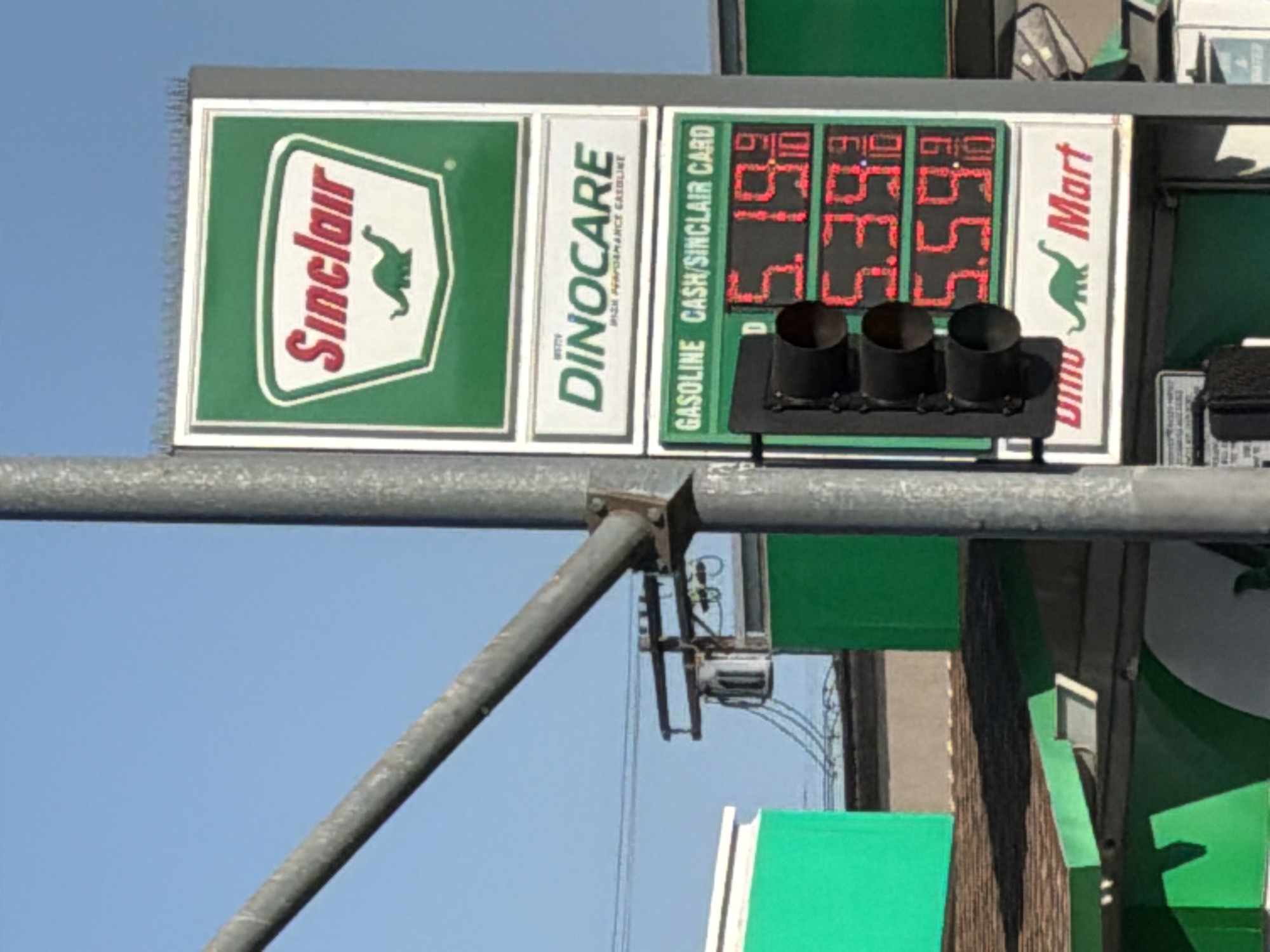 A gas station price sign is captured in an outdoor, daytime shot. The sign displays gas prices in red digital numbers against a green background. The prices are 5.19, 5.39, and 5.59. Above the prices, the sign indicates "GASOLINE CASH/SINCLAIR CARD". The sign also features the Sinclair logo, which includes a green dinosaur. Below the prices, the sign reads "Dino Mart" with a green dinosaur icon. The sign is mounted on a pole, and a traffic light is attached to the side of the sign. In the background, a clear blue sky is visible, along with parts of the gas station's buildings, which are painted green and brown. The overall scene suggests a typical gas station environment.